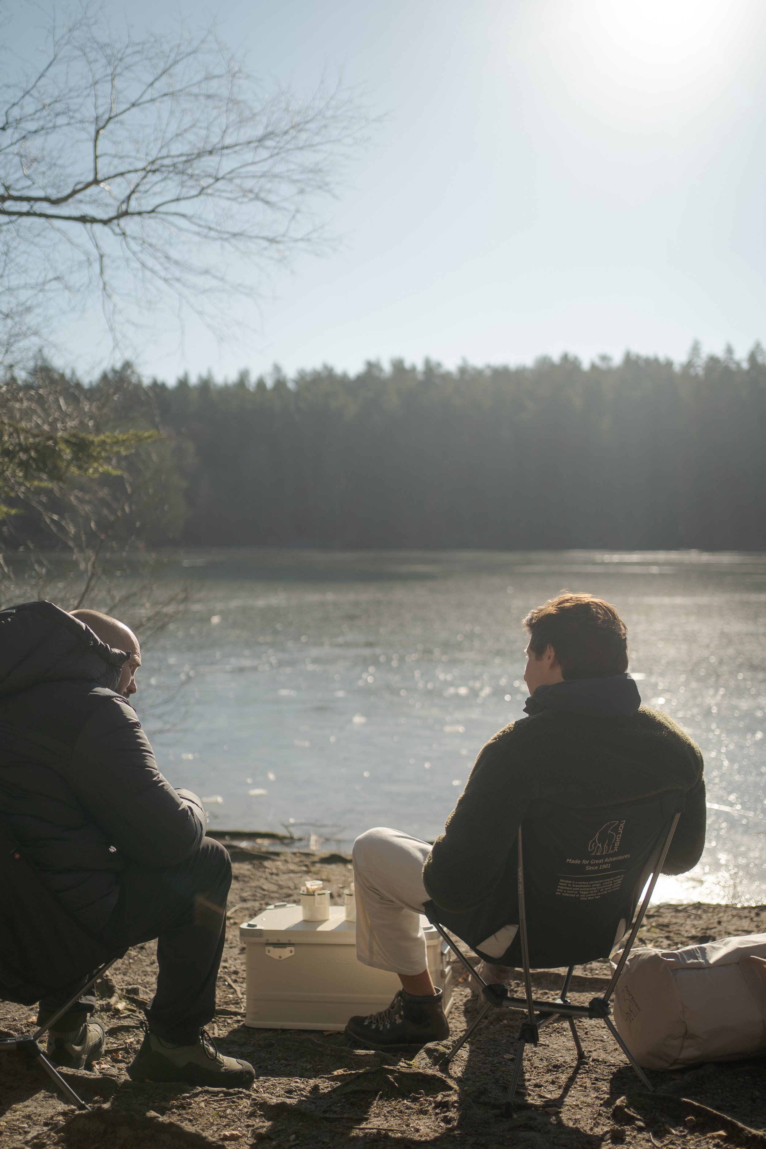 To personer sidder ved en sø med is, en person på en stol og en anden bøjet over, i naturen på en klar dag.