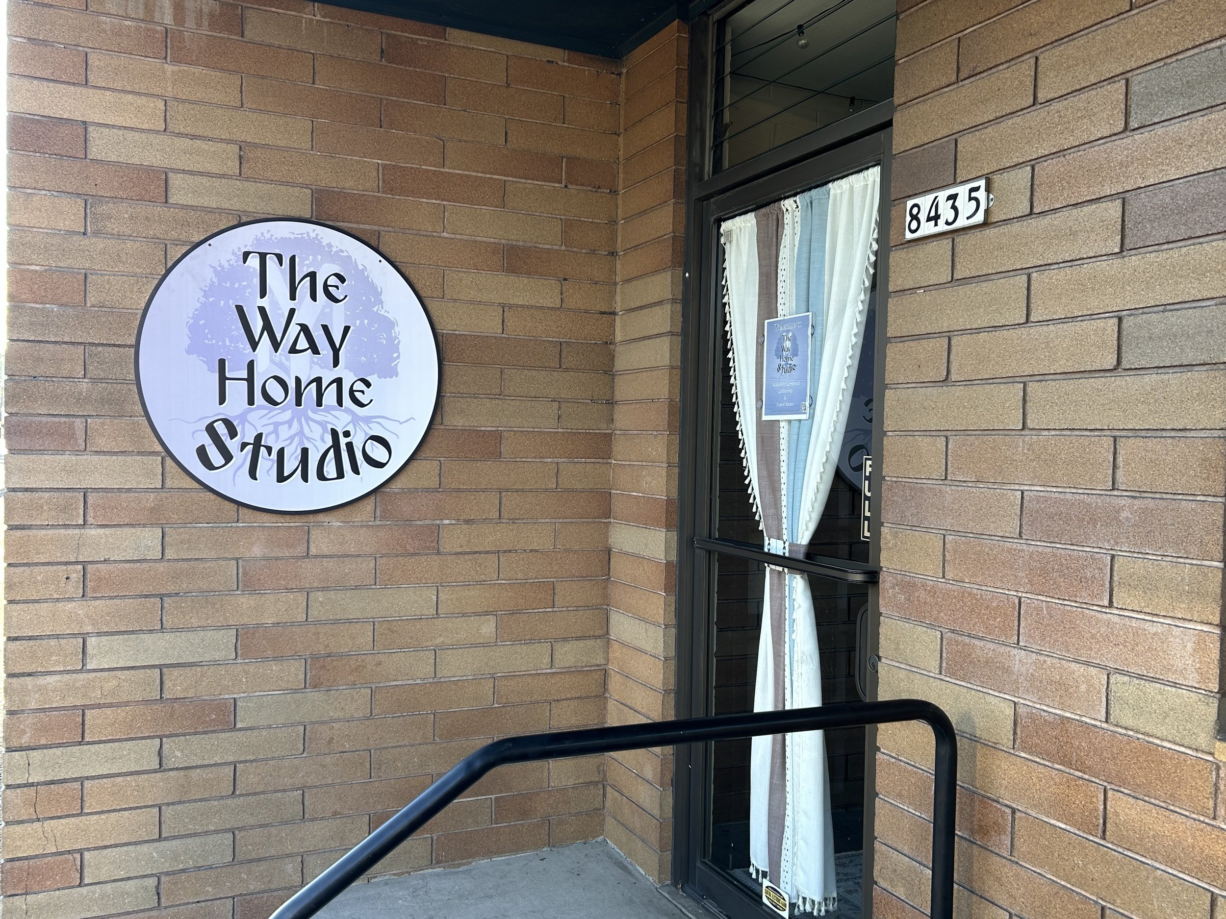 A storefront with a sign that reads 'The Way Home Studio' mounted on a brick wall. The entrance has a window with white, blue, and brown curtains and the street number 8435 above the window.