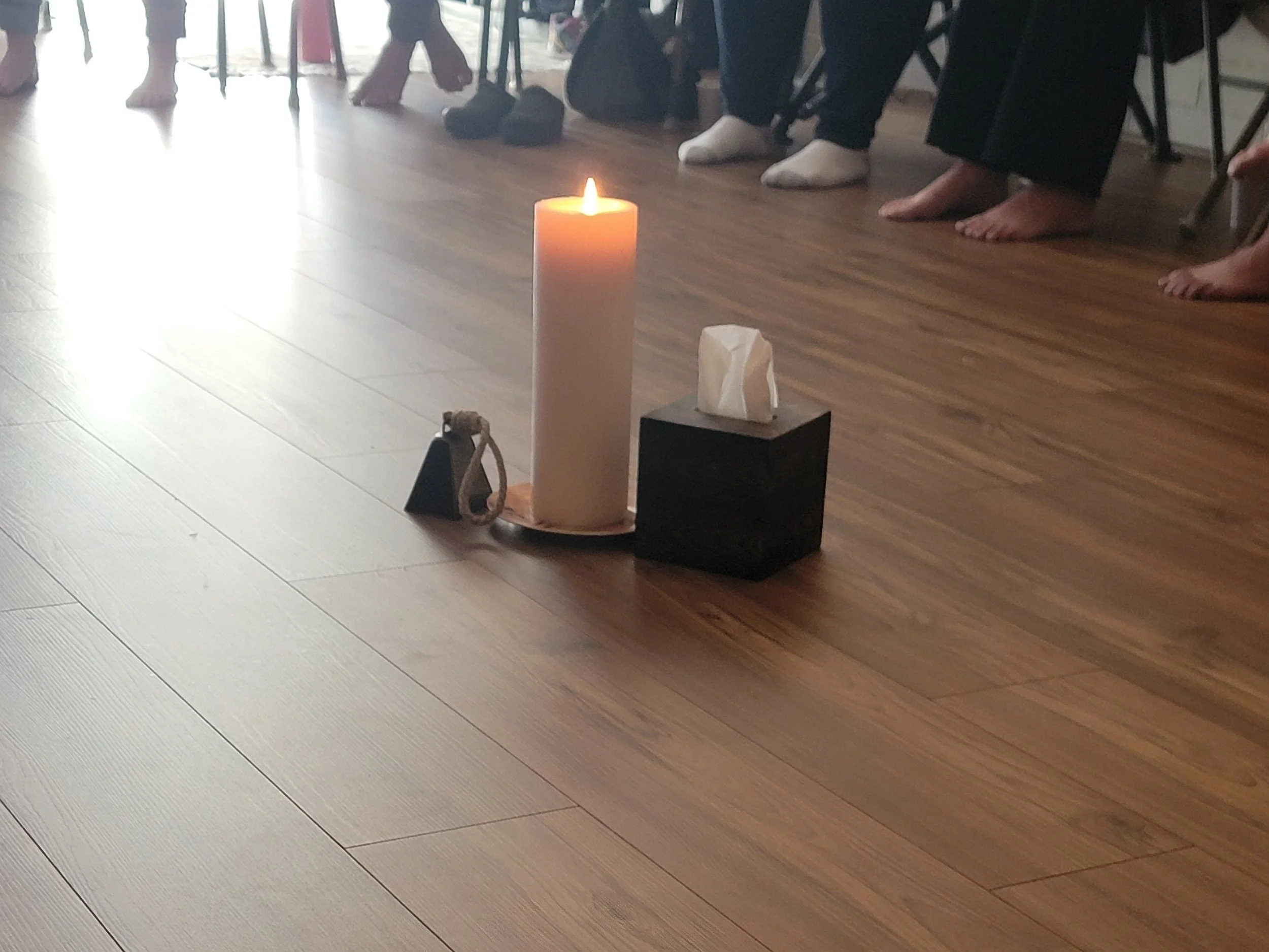 People standing barefoot on a wooden floor circle around a lit candle on a pedestal, possibly participating in a ritual or meditation.