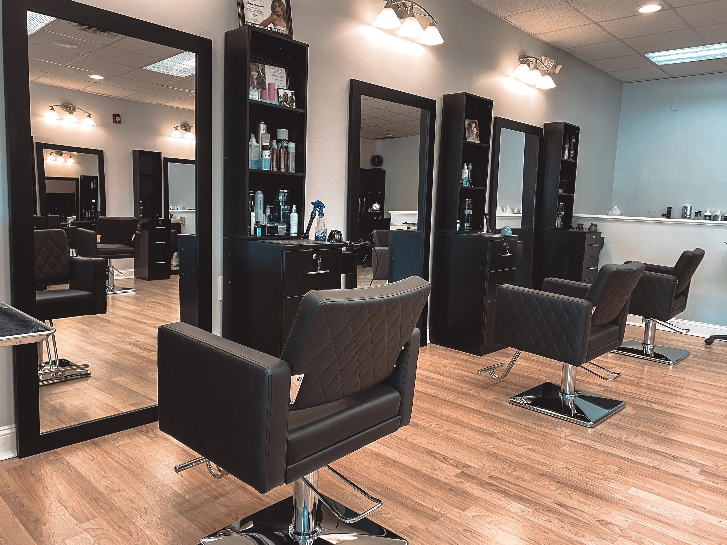 Empty hair salon with black styling chairs, large mirrors, black cabinets, wood flooring, and bright overhead lighting.