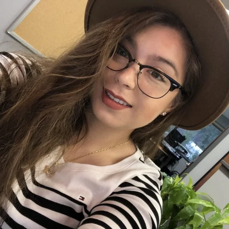 Young woman with glasses, long wavy hair, wearing a wide-brimmed hat, a striped shirt, and a necklace, smiling indoors with a plant and an office in the background.