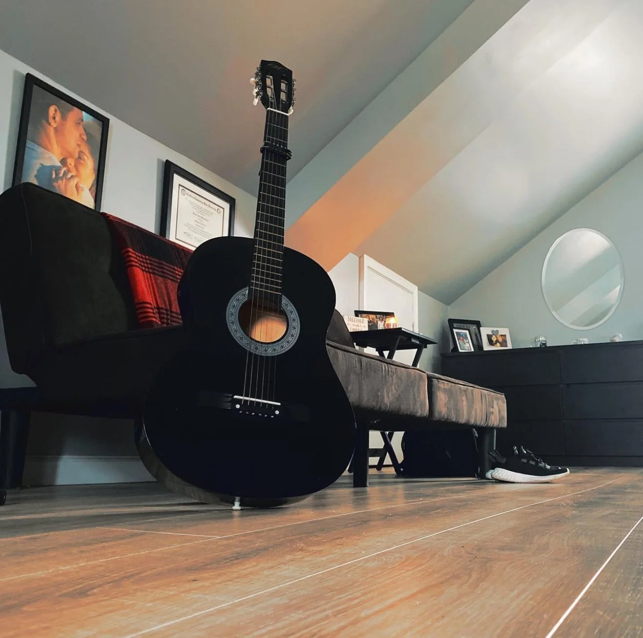 A black acoustic guitar leaning against a dark-colored sofa in a room with wooden flooring and slanted ceiling. On the wall, there are framed pictures and certificates, including a portrait of two children.