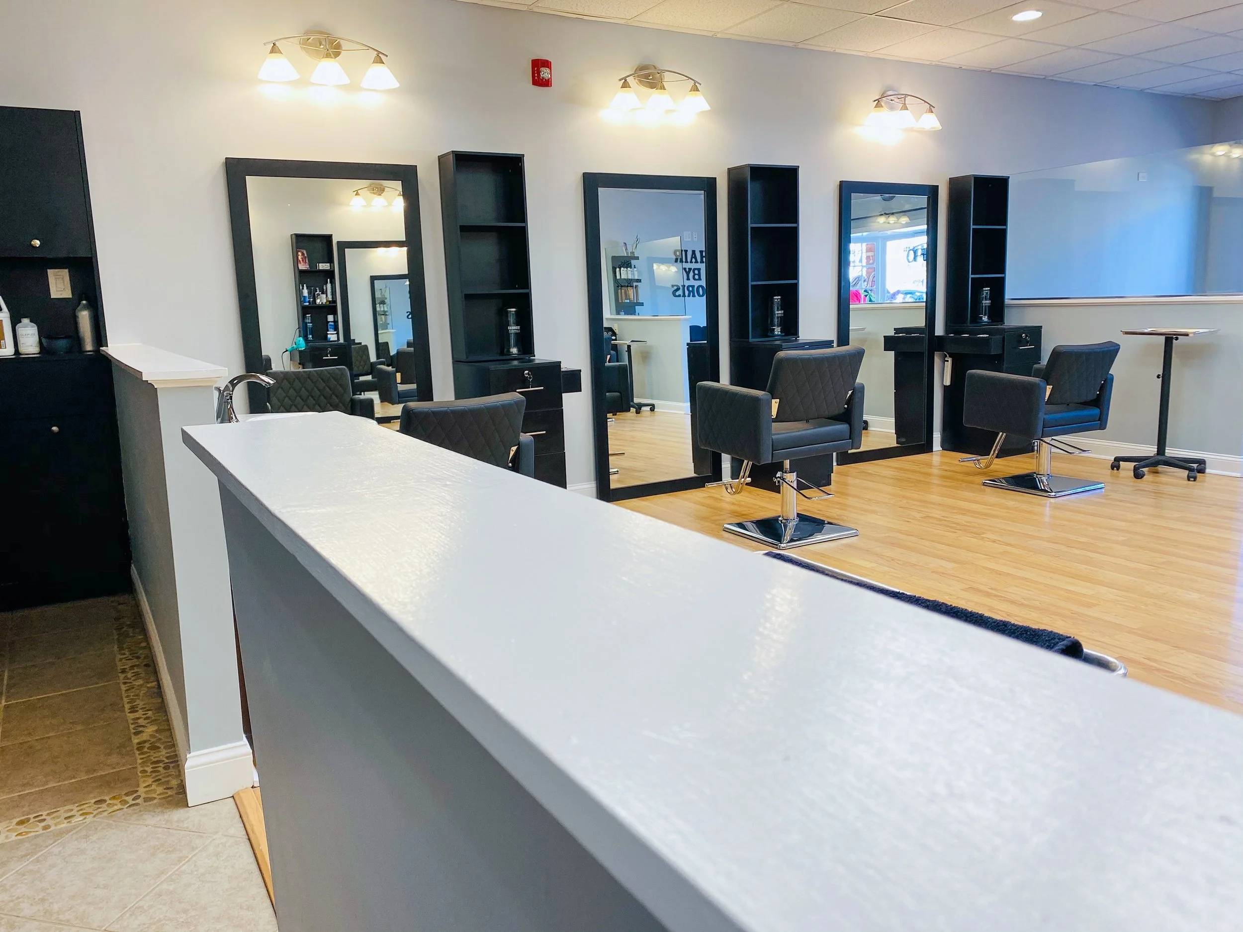 A modern hair salon with four styling stations, each with a large mirror and black salon chairs, hardwood floors, and bright lighting.