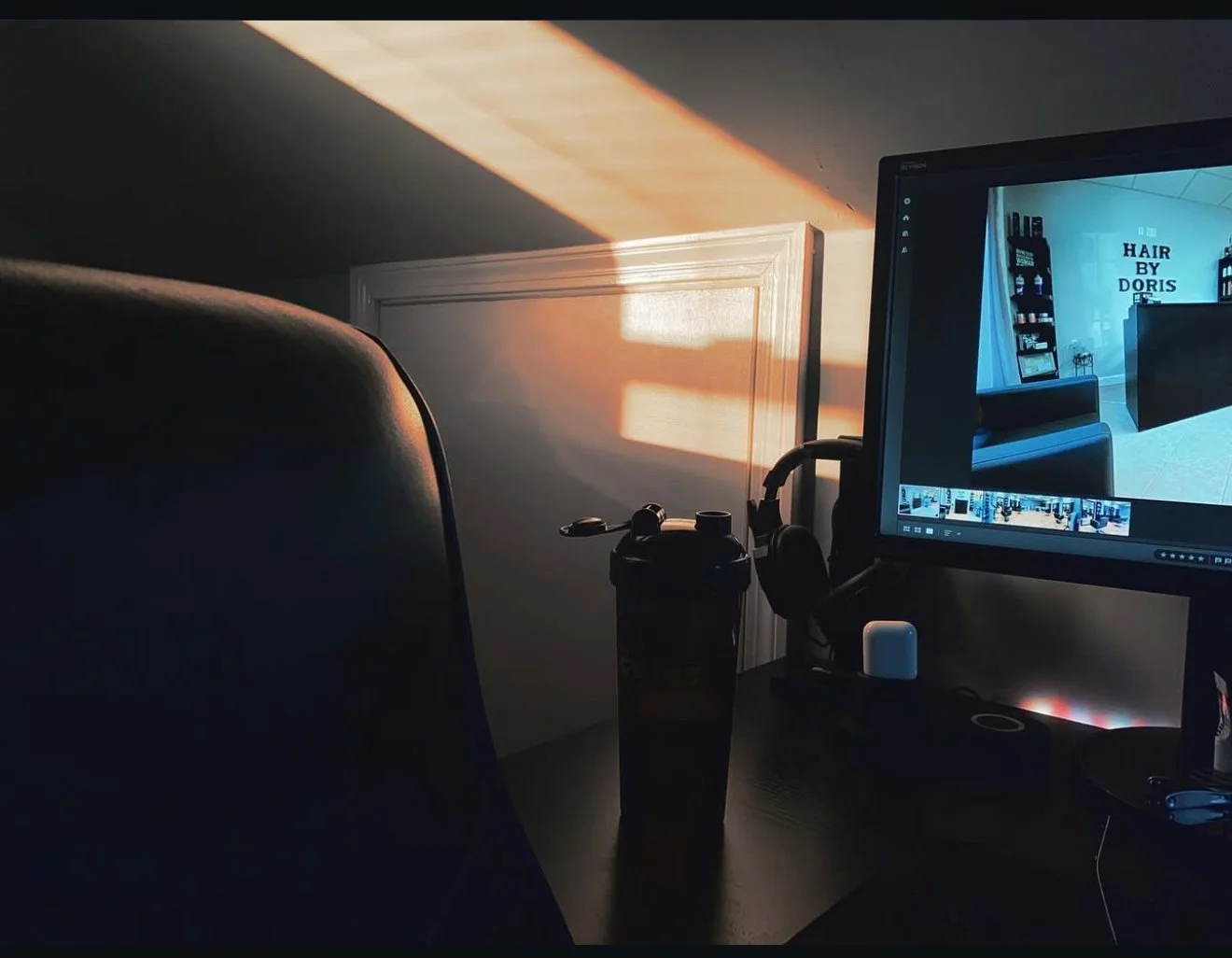 A computer desk setup showing a monitor with a photo editing window, a dark water bottle, and a partial chair, with sunlight casting a warm glow on the wall behind.