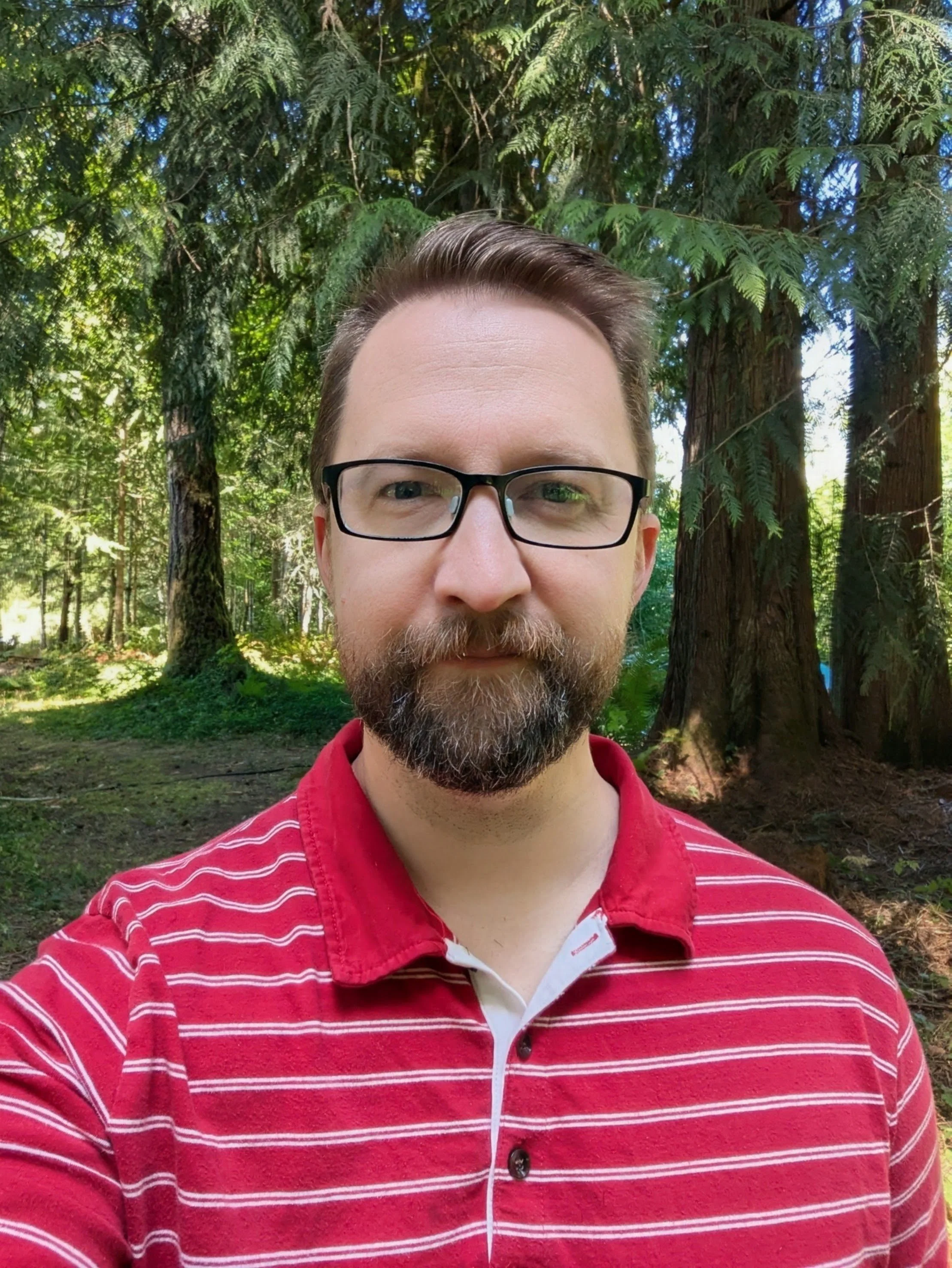 A man taking a selfie outdoors surrounded by tall trees with lush green foliage. He is wearing glasses and a red and white striped polo shirt.