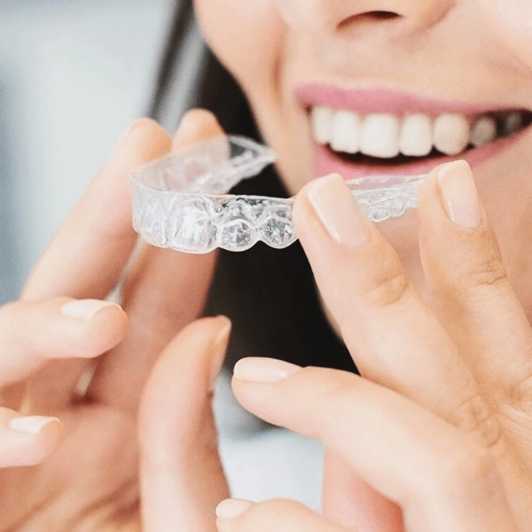 A woman holding a clear Invisalign dental aligner near her teeth and smiling.