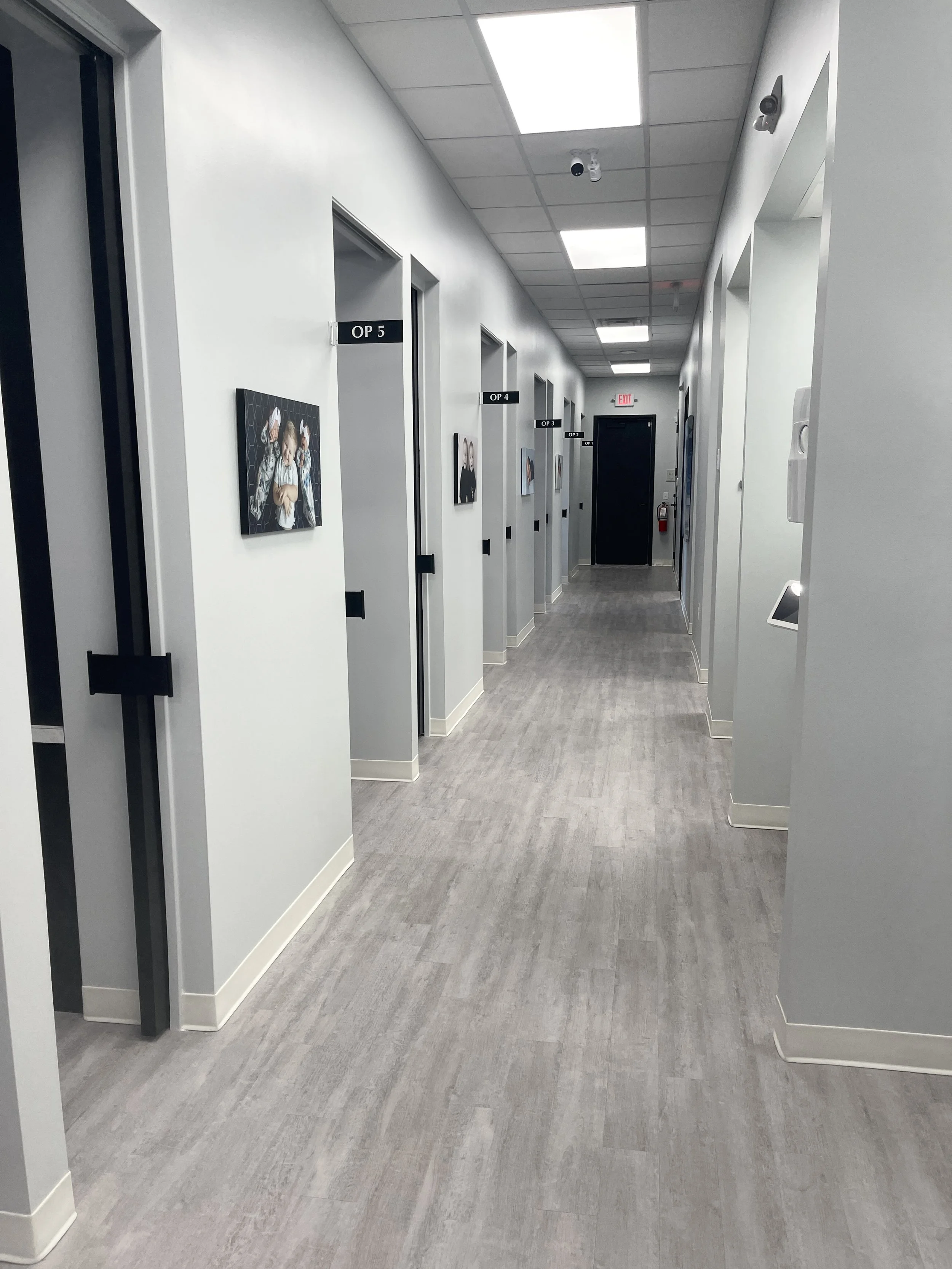Empty hallway in a medical or office facility with numbered doorways, framed photographs on the wall, overhead fluorescent lighting, and a security camera.