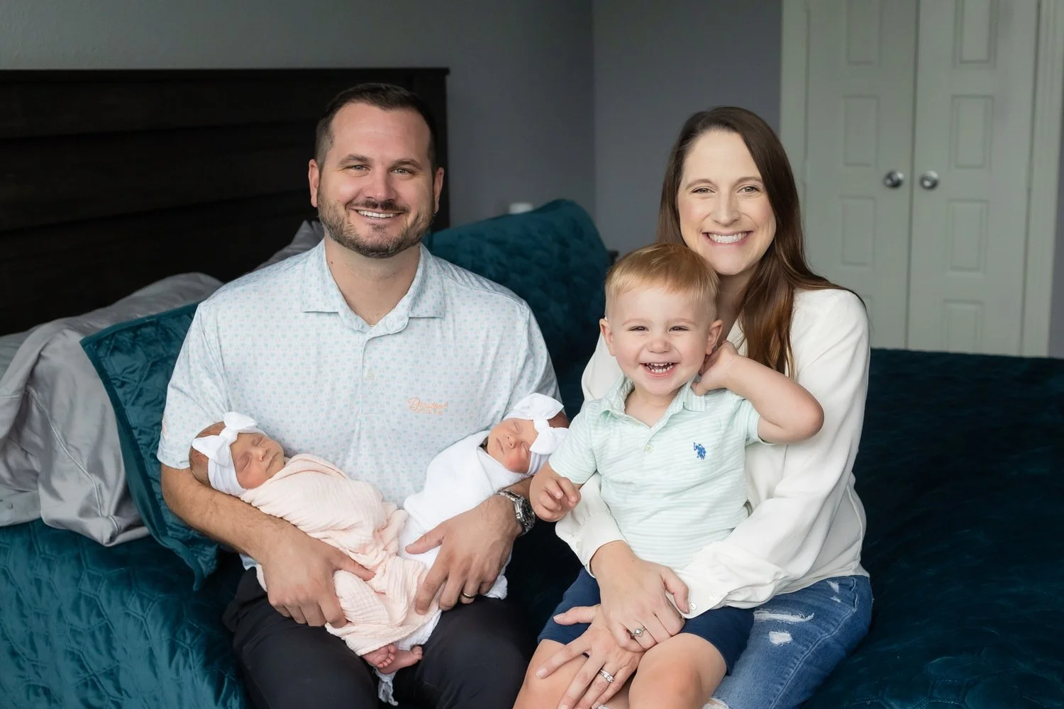 A family of four sitting on a bed, with two newborn babies and a toddler, smiling at the camera in a bedroom.