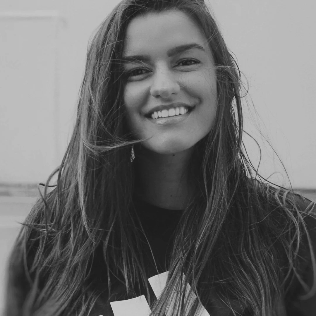 Black-and-white photo of a young woman with long, wavy hair, smiling and showing her teeth, wearing a dark shirt with a white graphic design.