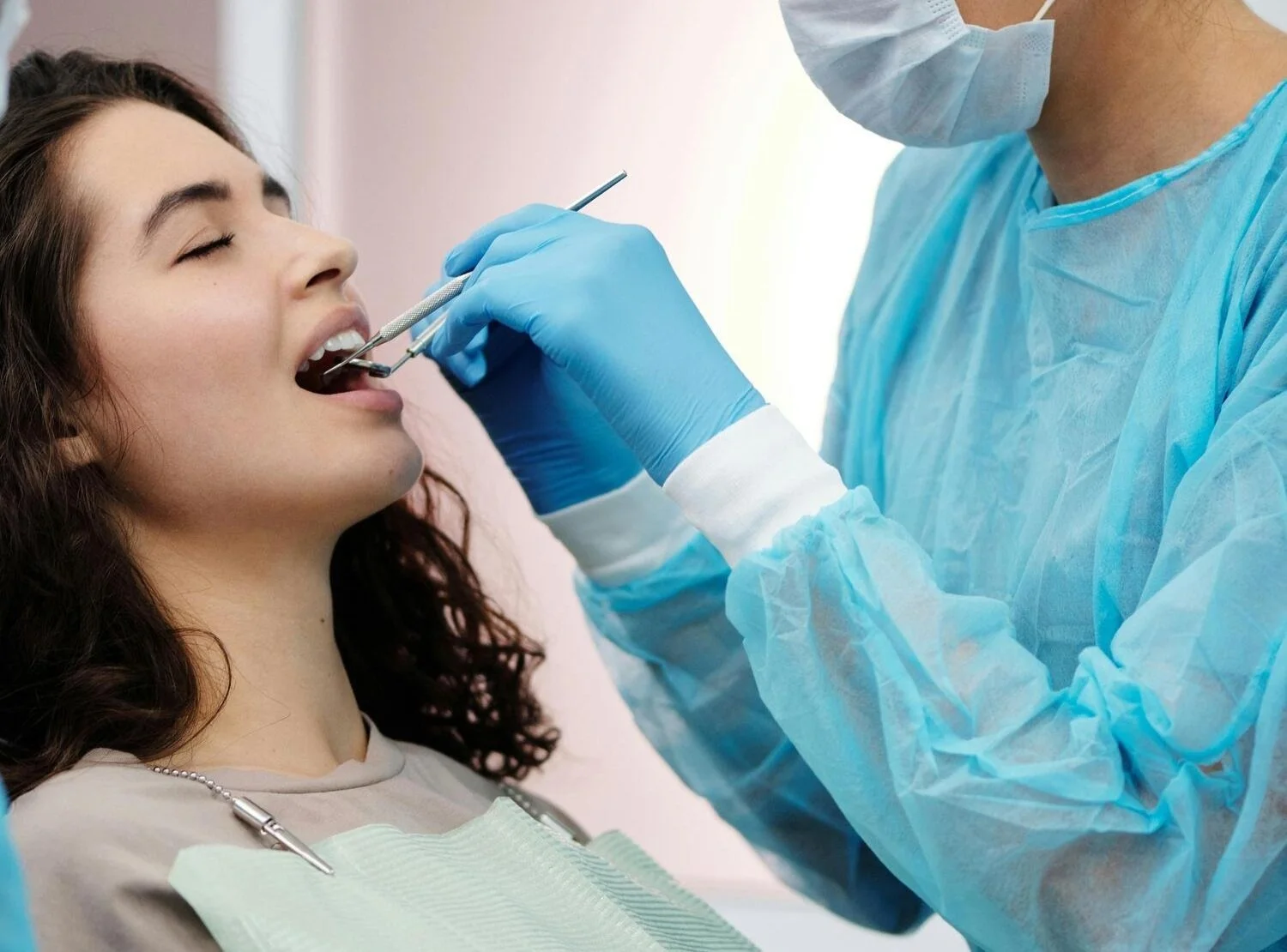 A woman in a dental chair with her mouth open, receiving a dental examination or procedure from a dentist wearing blue gloves, a mask, and a gown.