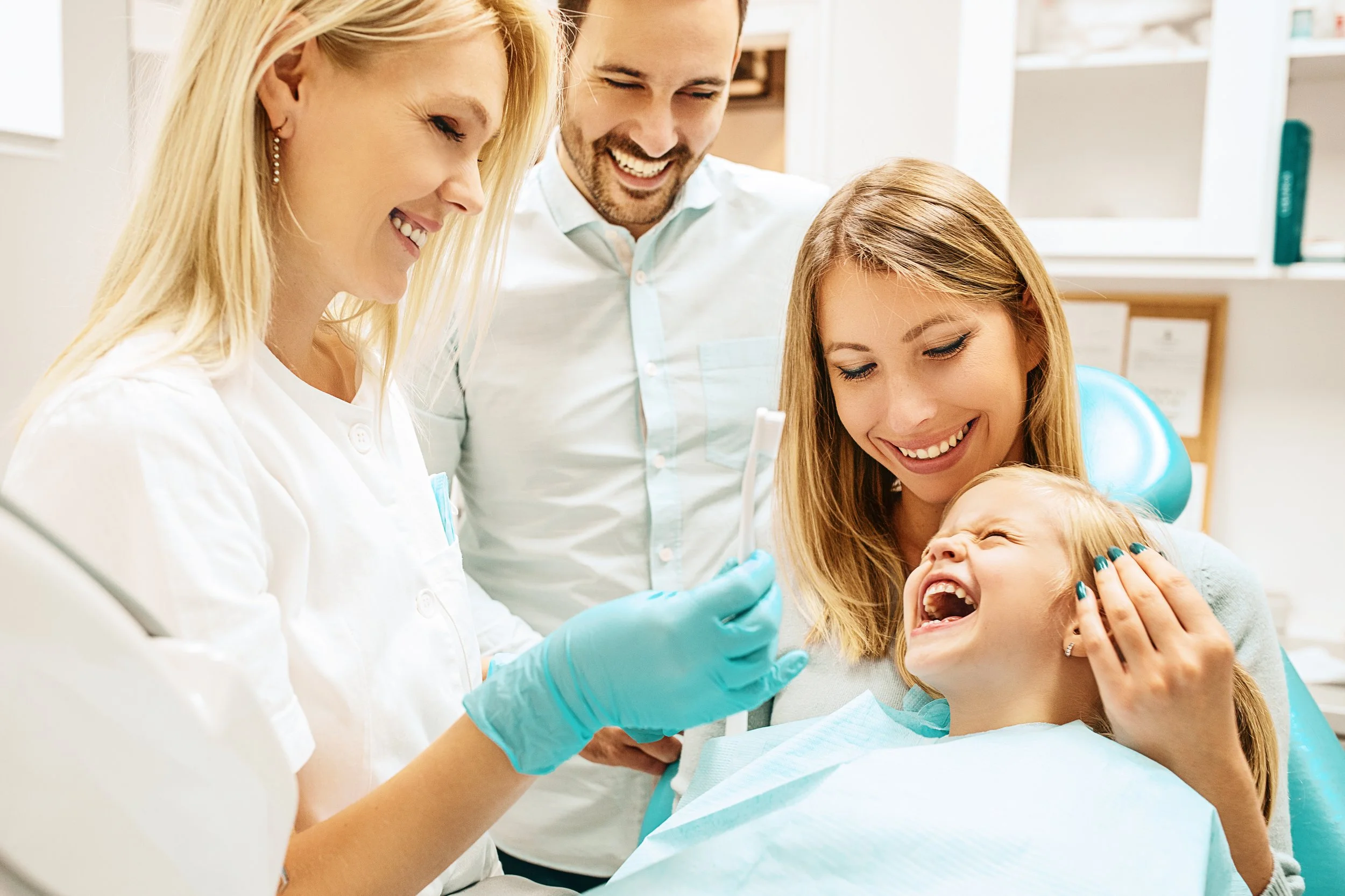 A young girl laughing during a dental checkup with a woman holding her head and dental staff showing her a toothbrush.
