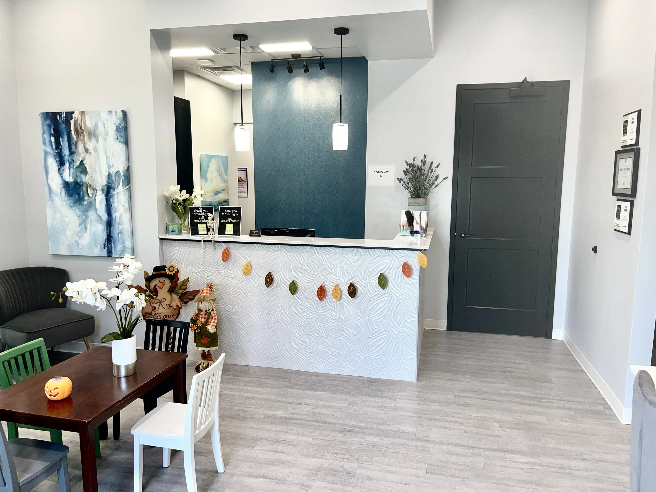 Reception desk in a modern office or waiting area decorated with autumn leaves garland, a snowman figure, and fall-themed decorations. There are paintings on the wall, a vase with white flowers on the desk, and a small pumpkin candle on a table with chairs nearby.