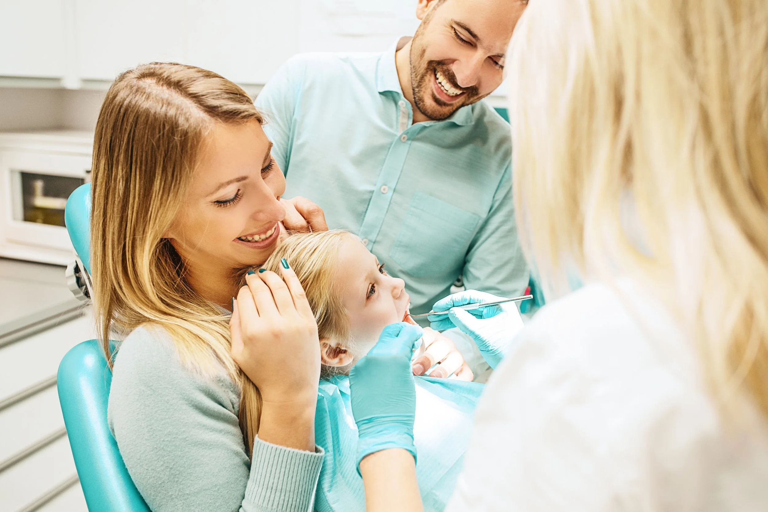 A young girl sitting in a dental chair, with her mother holding her, as a dentist examines her mouth while the father watches and smiles.