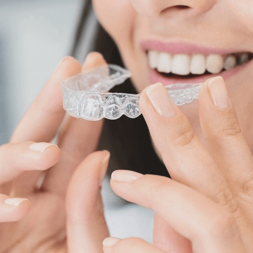 Close-up of a smiling woman holding a clear dental aligner near her teeth.