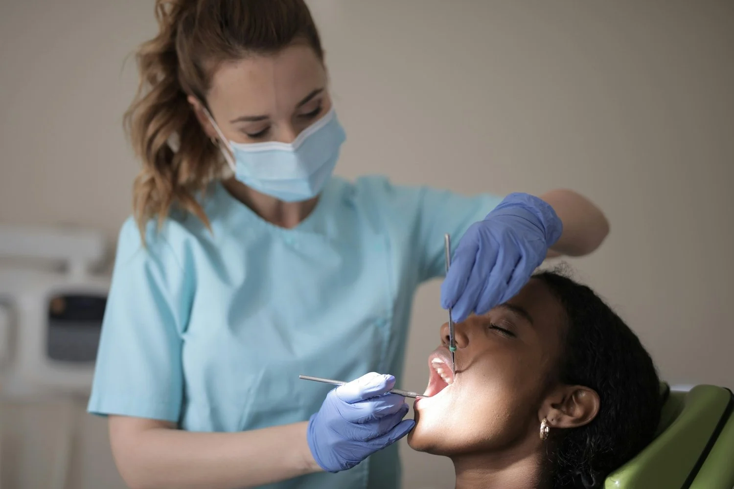 Dental professional wearing a face mask and gloves examining a woman's mouth with dental tools in a clinic.