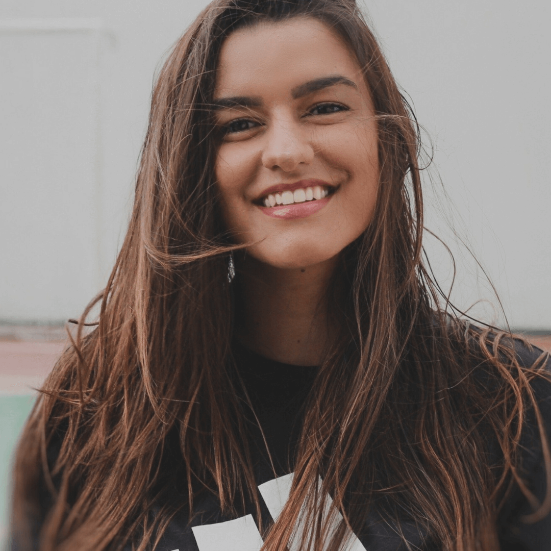 Close-up of a smiling woman with long brown hair and white teeth, wearing a black shirt, outdoors with a blurred background.