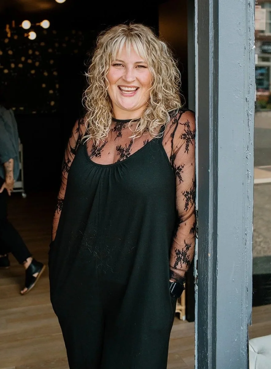 A woman with blonde curly hair, wearing a black dress with a bow, standing outside a shop with a large window, smiling and posing.