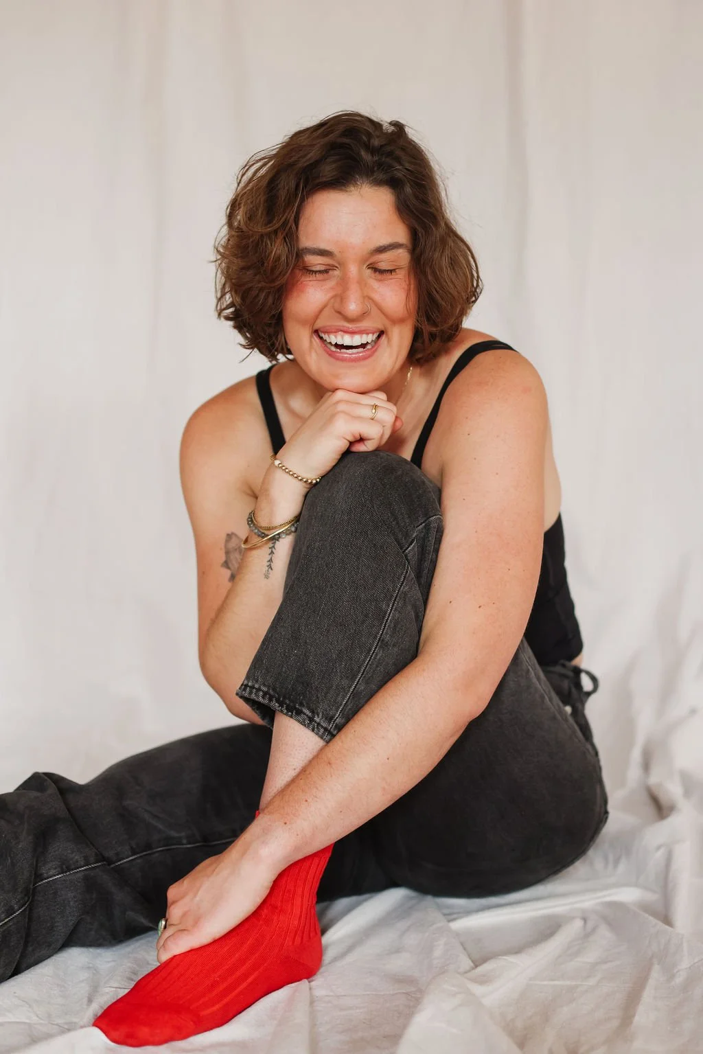 A young woman with short, wavy brown hair, wearing a black sleeveless top and dark jeans, sitting on a light-colored fabric surface, smiling with eyes closed, wearing red socks, and holding her left foot with her right hand.