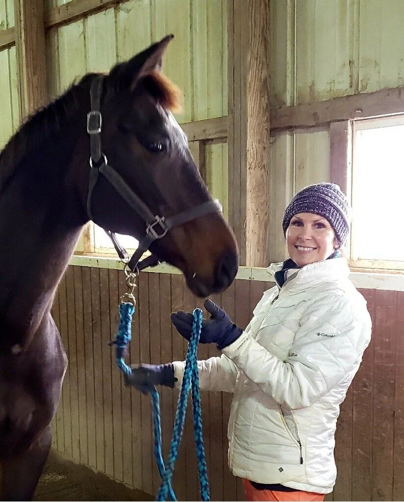 A woman with a knit cap and white jacket smiling while holding a blue lead rope attached to a dark brown horse inside a barn with wooden and green walls, and a window letting in natural light.