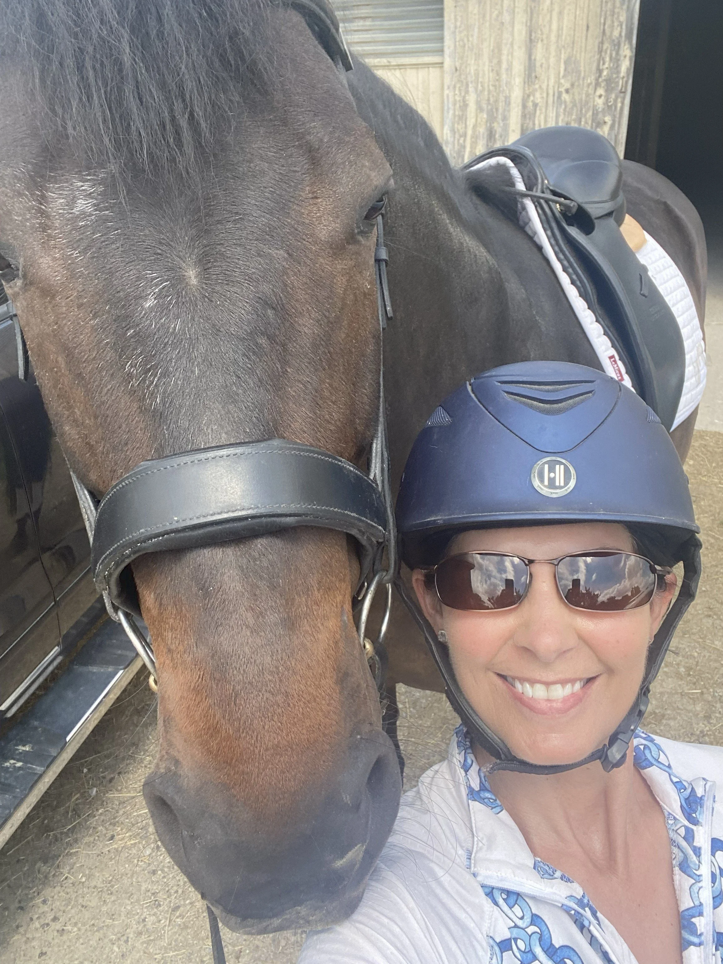 A woman wearing a helmet and sunglasses taking a selfie with a brown horse.