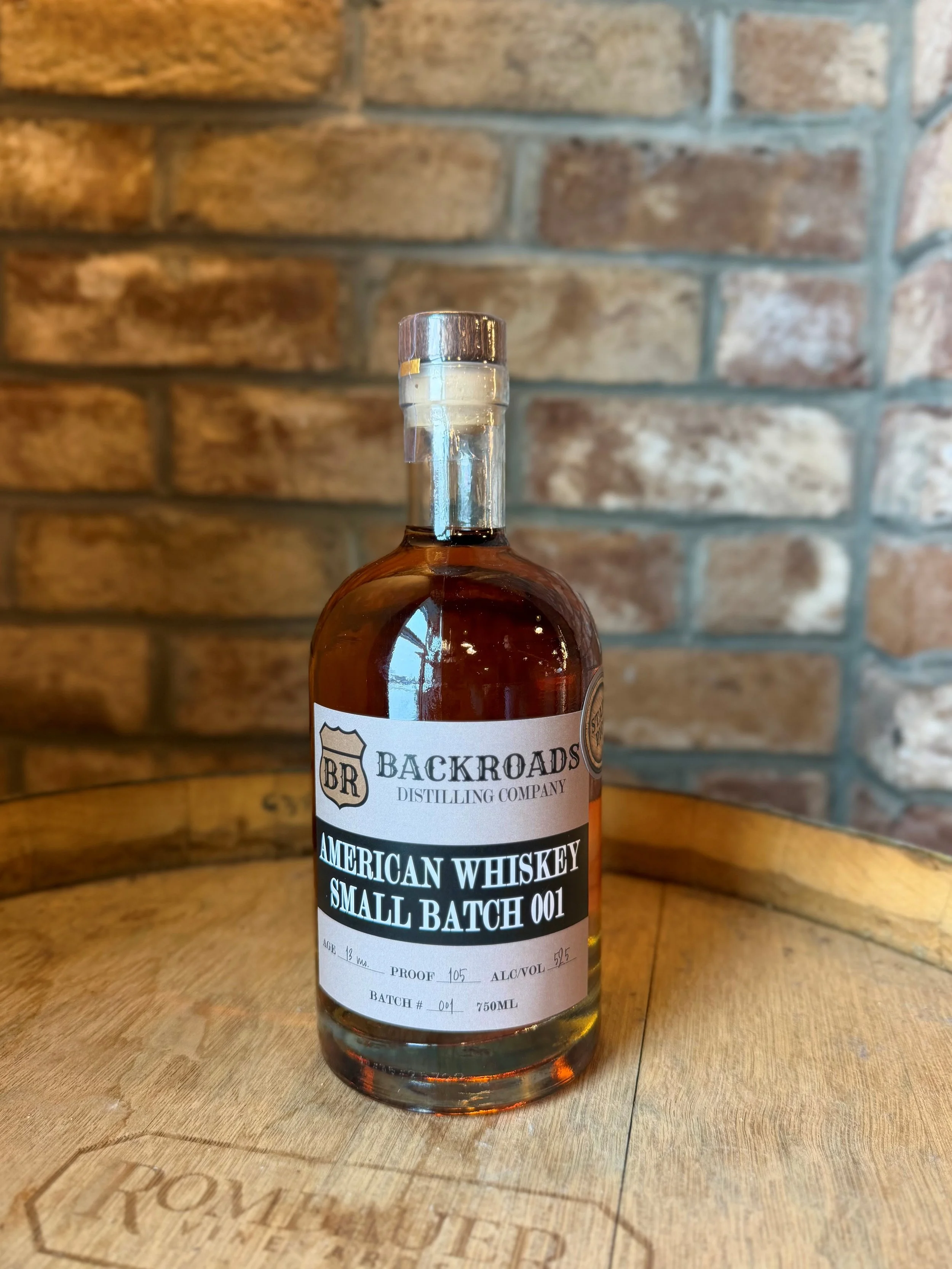 A bottle of Backroads Distilling Company American Whiskey Small Batch 001 on a wooden surface with a brick wall background.
