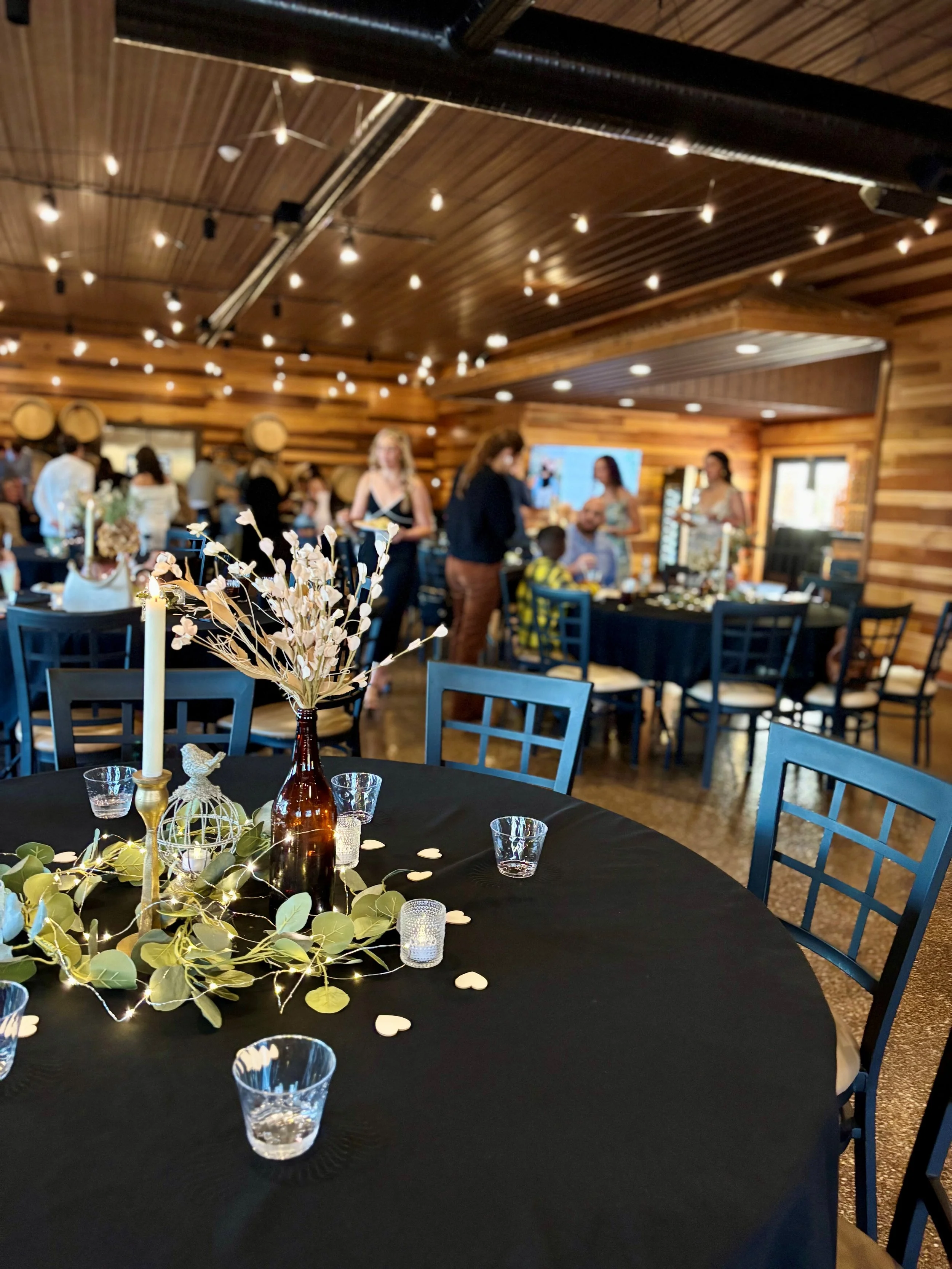 Decorated banquet table with a black tablecloth, white candles, a brown bottle with dried flowers, string lights, and small candles or tealights, at a rustic wooden event venue with people mingling in the background.