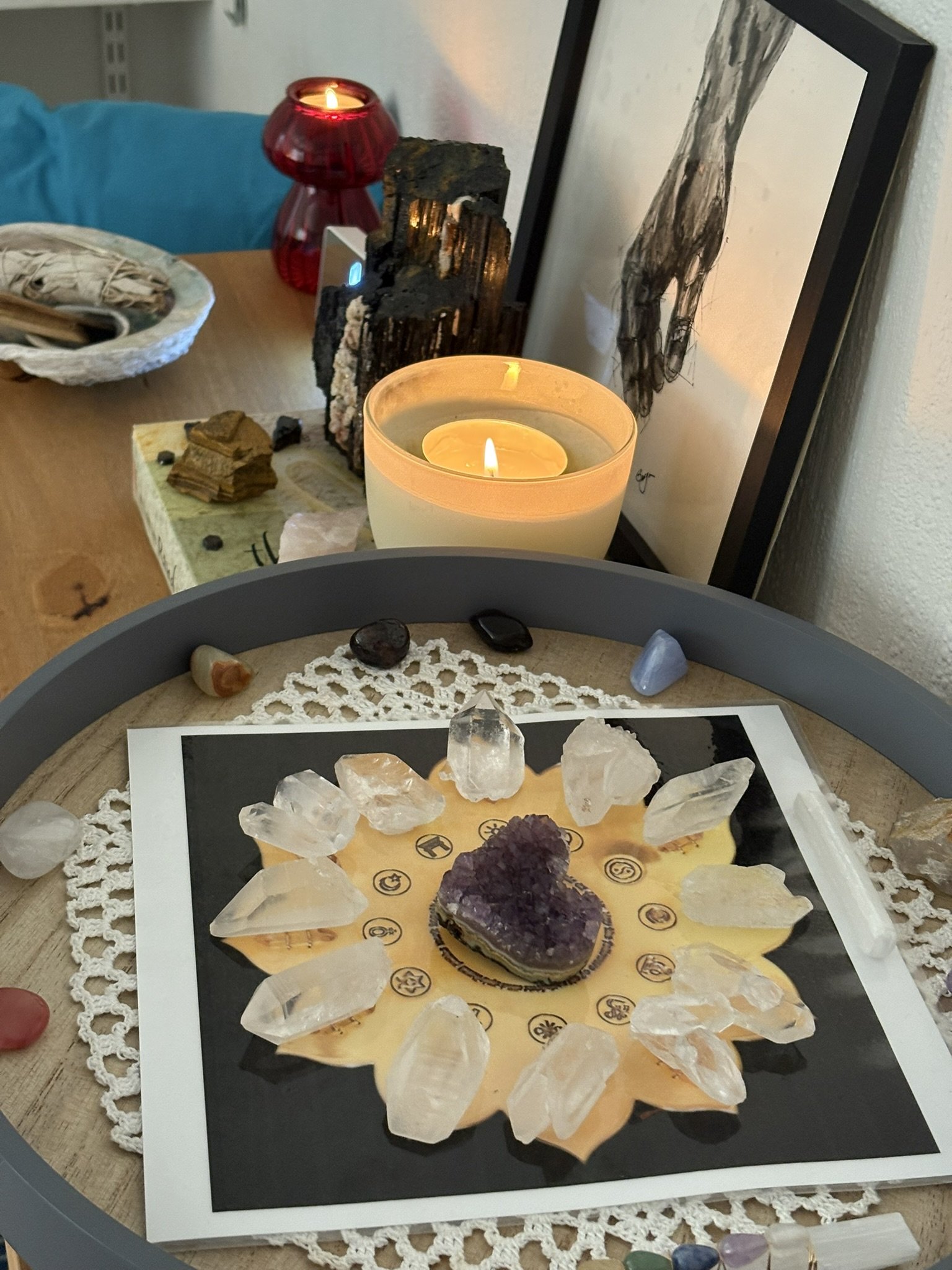 Clear quartz crystals, amethyst, and other small gemstones in a circle on a crystal grid , with a purple amethyst cluster in the centre, on a wooden table. In the background is a burning candle, framed artwork, and a red candle holder.