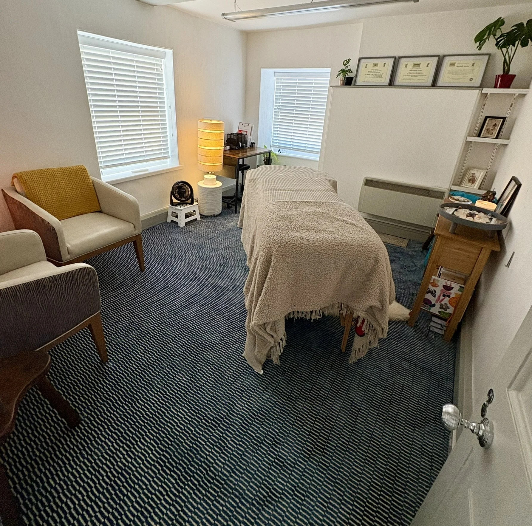 A Reiki Therapy room with a massage table covered with a blanket, a small wooden table, three framed certificates on a white shelf, a lamp, a small desk, and two armchairs in a room with white walls, window blinds, and carpet.