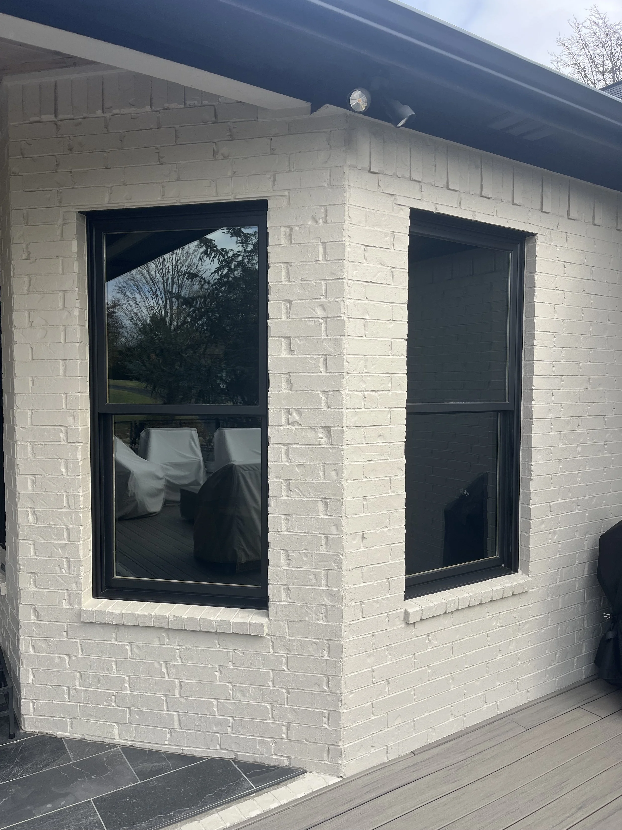 Exterior corner of a house with white brick walls, two black-framed windows, a roof with grey eaves, and a tiled porch area.