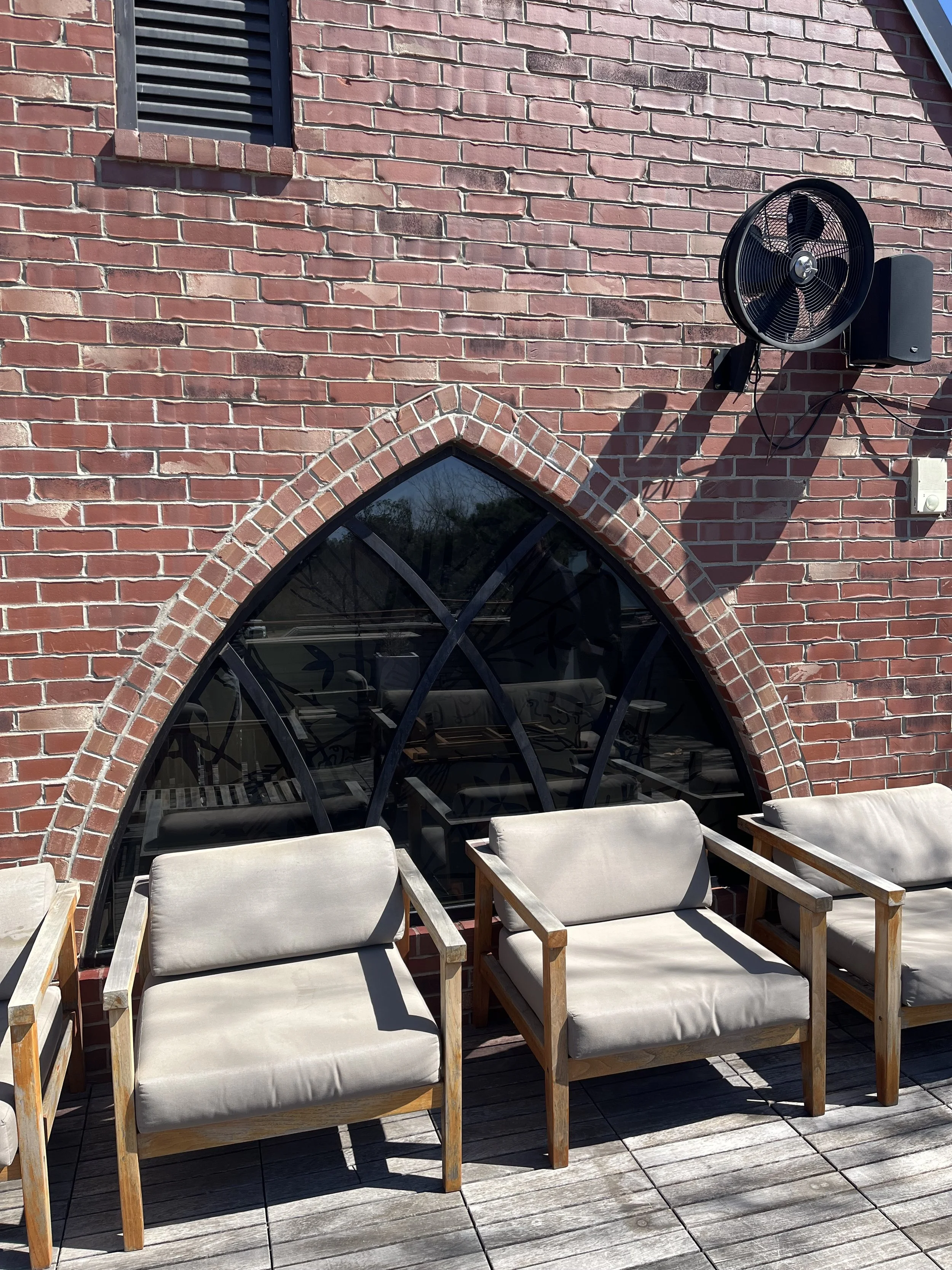 Outdoor seating with chairs in front of a brick building with arched window, wall-mounted fan, and speaker.