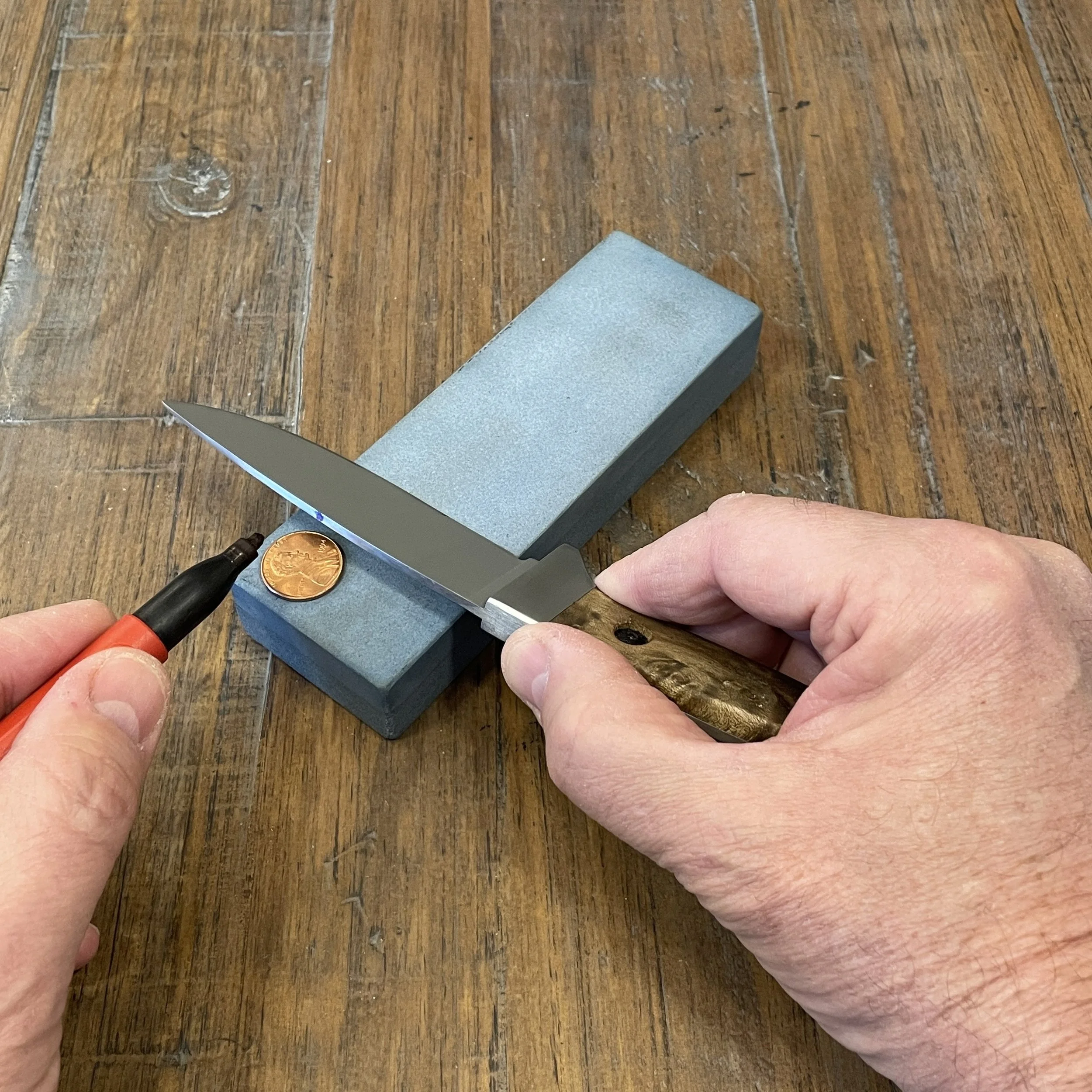 A knife blade and a penny resting on a sharpening stone.  A dot is being applied to the spine of the blade with a permanent marker in preparation for determining the most recent sharpening angle using a coin stack for reference.