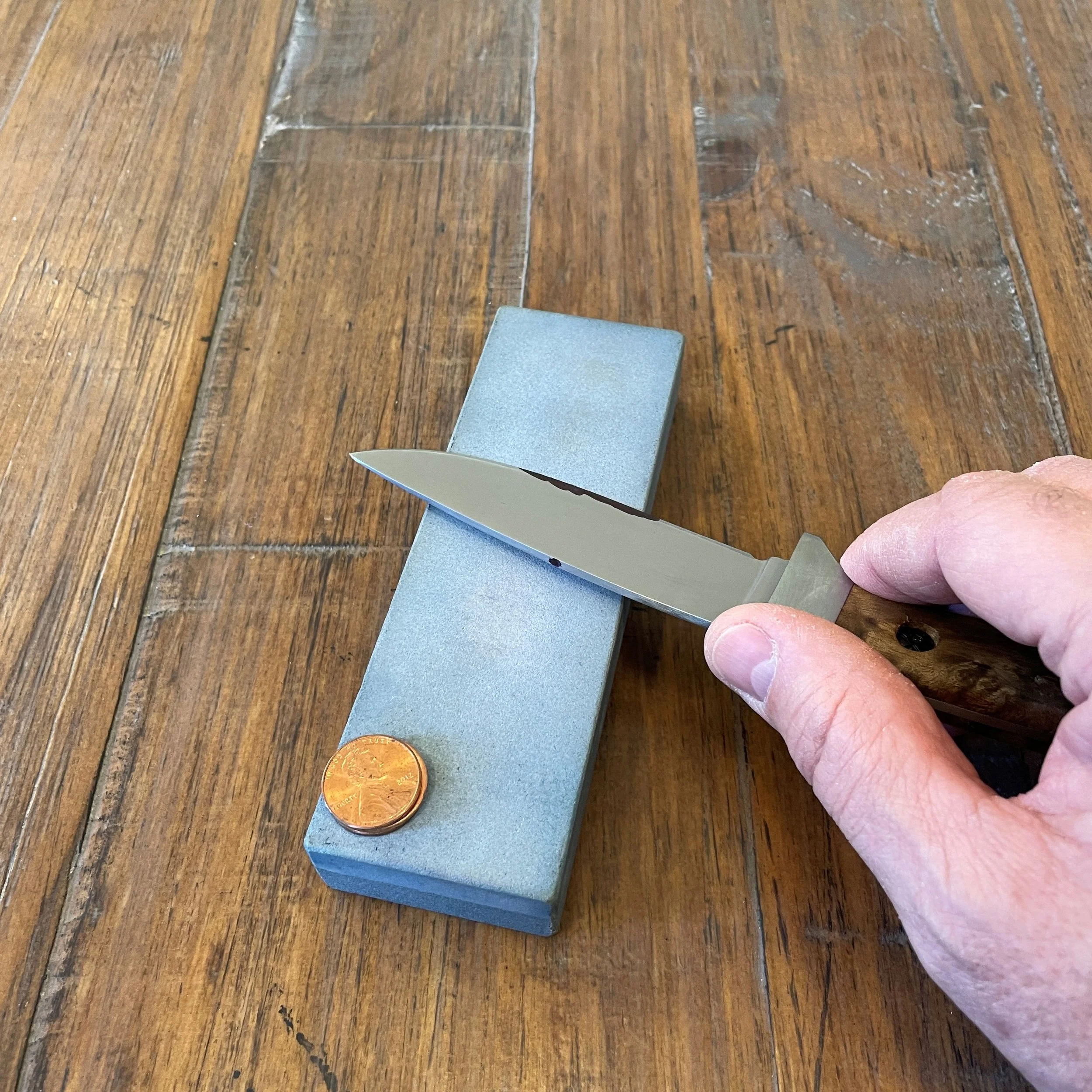 A hand holding a knife against the middle of a sharpening stone with a penny resting on the near end of the stone.  This is an active sharpening stroke to determine the most recent sharpening angle using a coin stack for reference.