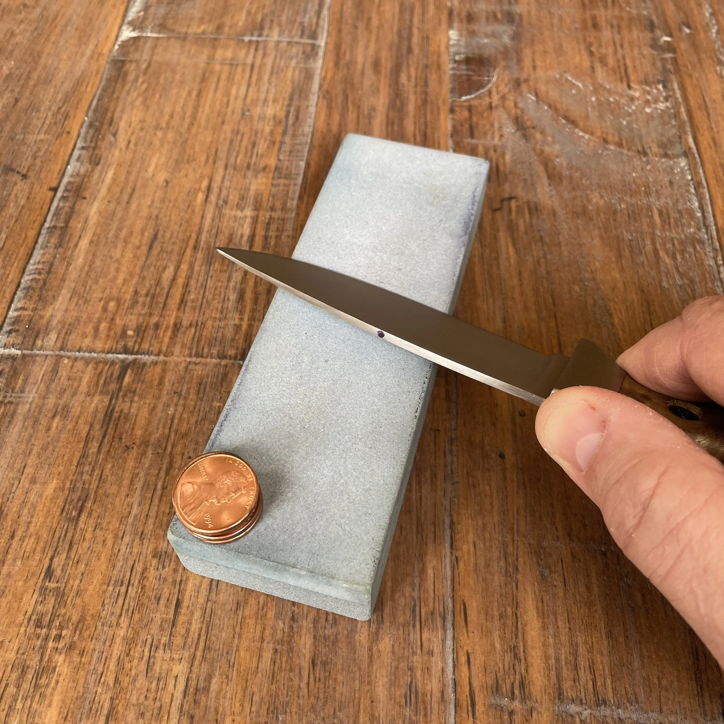 A hand holding a knife against the middle of a sharpening stone with a penny resting on the near end of the stone. This is an active sharpening stroke when sharpening to a specific desired angle using a coin stack for reference.