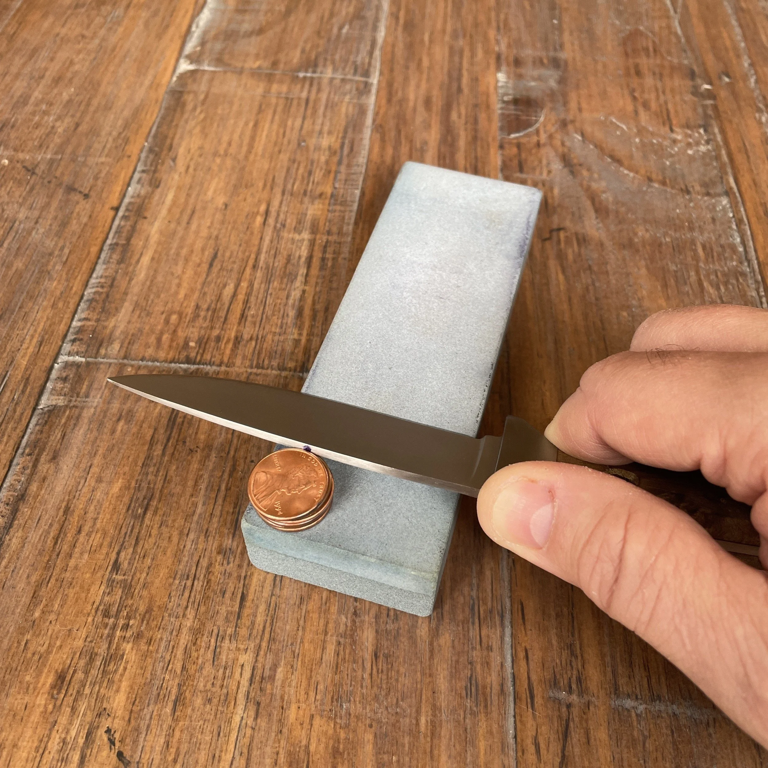 A hand holding a knife against a sharpening stone with the spine aligned with a penny resting on the stone. This is the beginning of a sharpening stroke when sharpening to a specific desired angle using a coin stack for reference.
