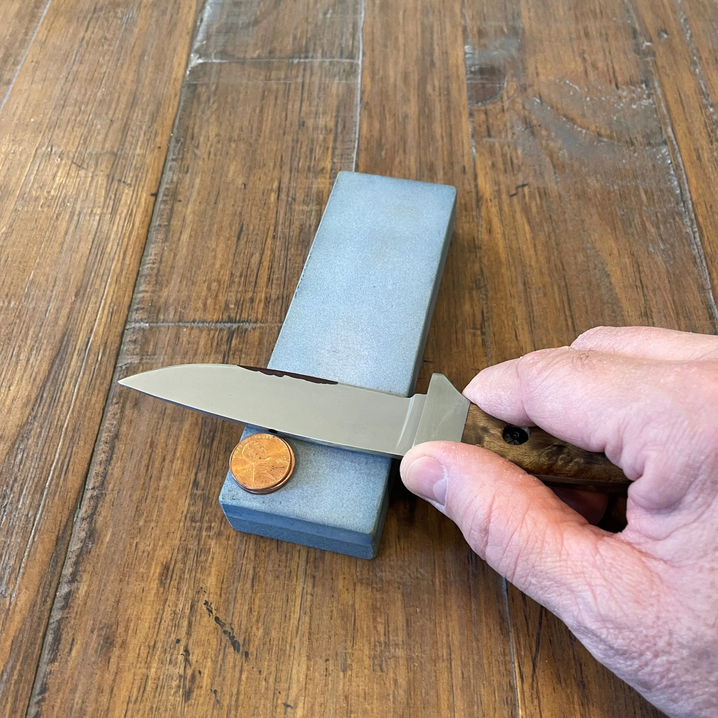 A hand holding a knife against a sharpening stone with the spine aligned with a penny resting on the stone.  This is the beginning of a sharpening stroke to determine the most recent sharpening angle using a coin stack for reference.