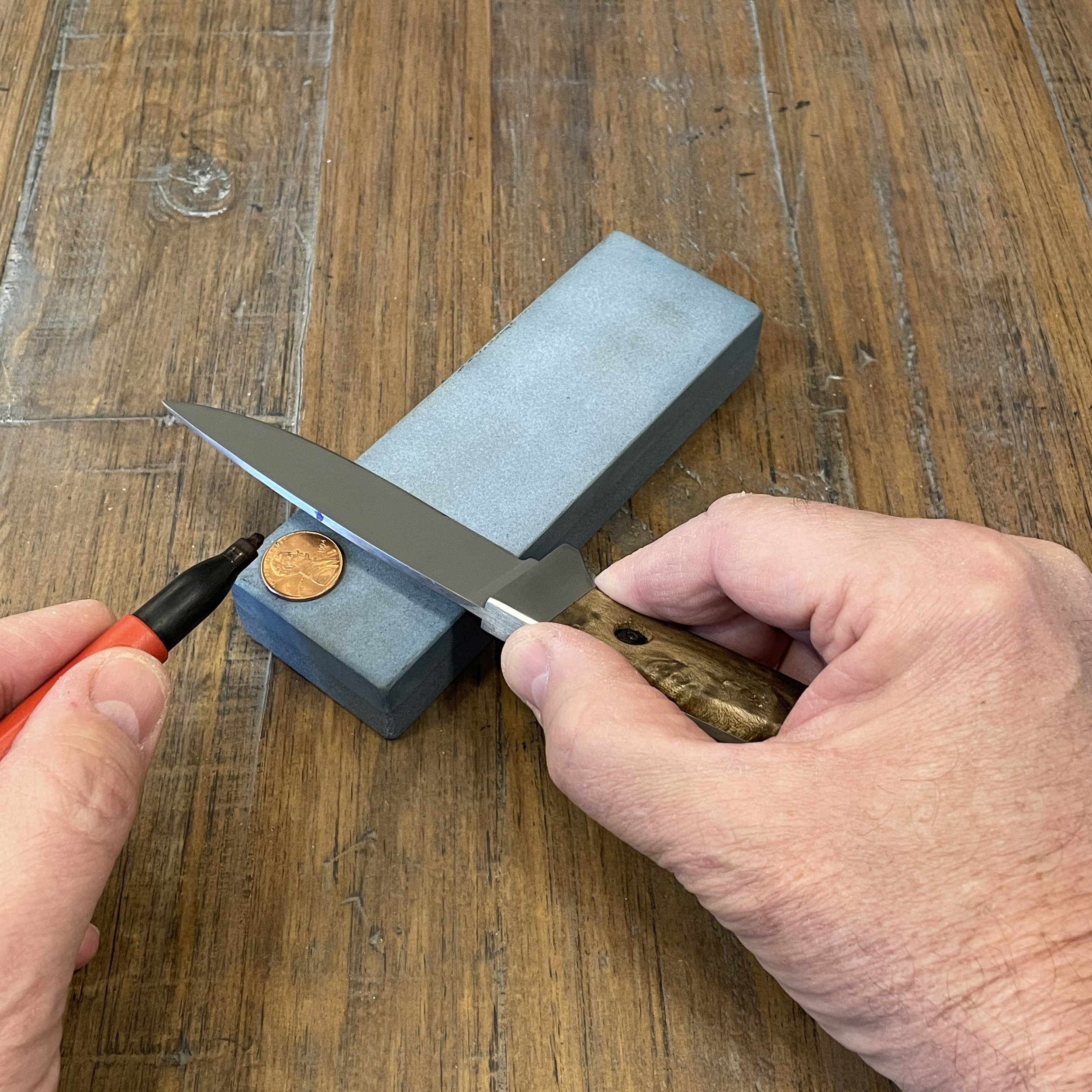 A knife blade and a penny resting on a sharpening stone. A dot is being applied to the spine of the blade with a permanent marker in preparation for sharpening the blade to a desired angle using a coin stack for reference.