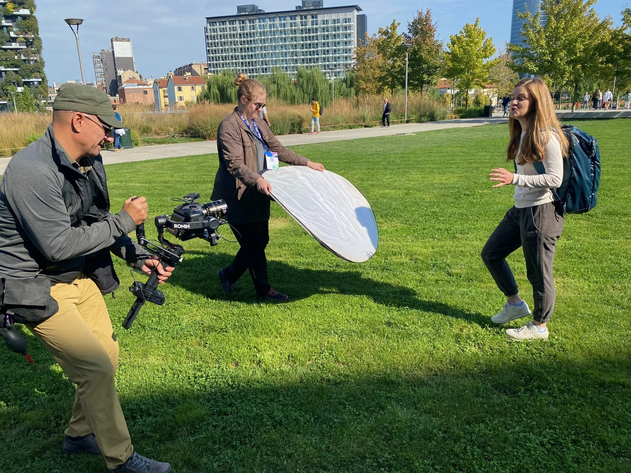 A young woman with long hair, wearing a white sweater and gray pants, stands on grass with a backpack, speaking to a camera crew. A female crew member holds a reflector, and a male crew member operates a camera mounted on a stabilizer.