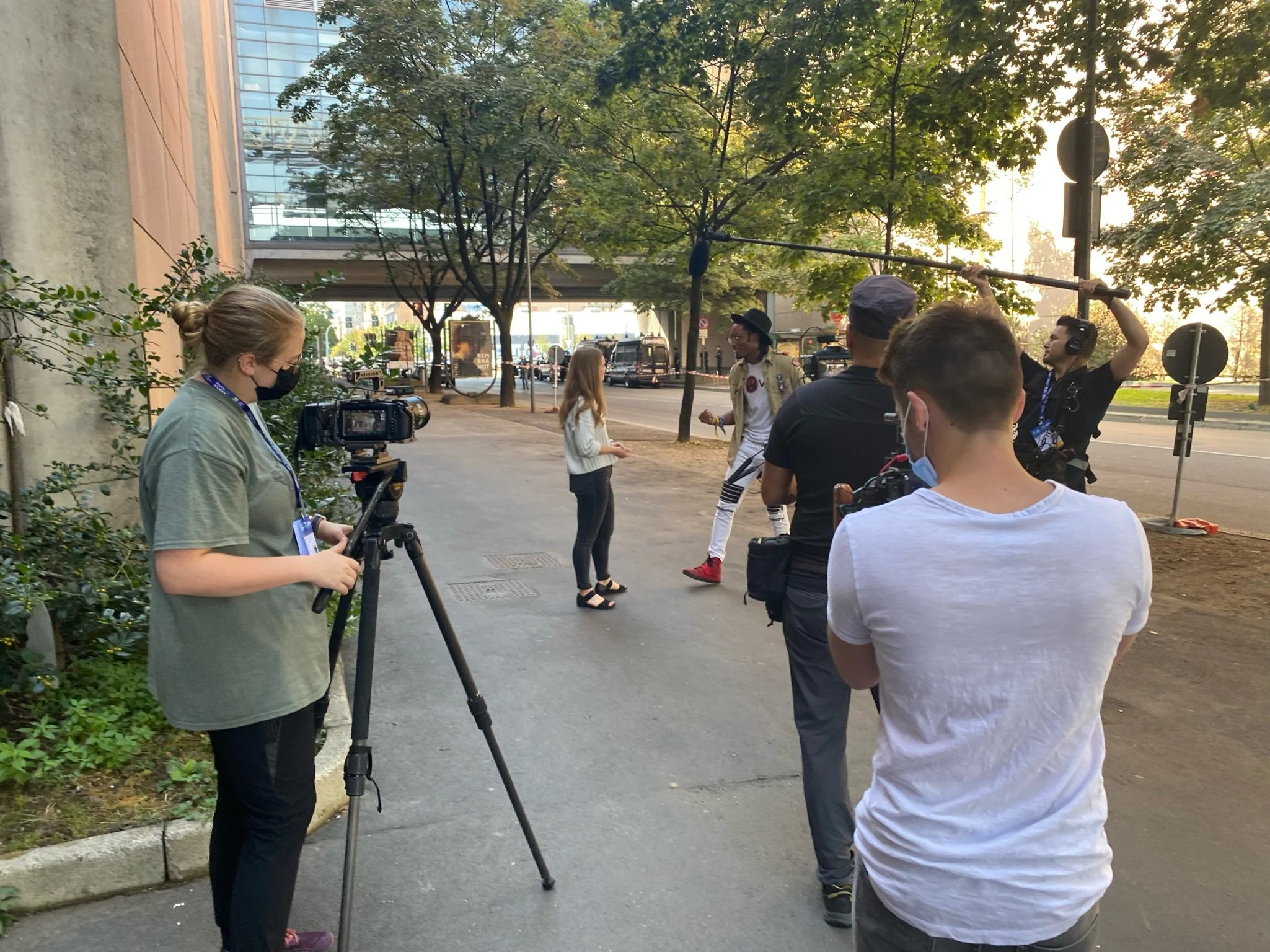 Film crew filming a scene on a city sidewalk with trees and modern buildings in the background. Crew members include a woman with a camera on tripod, a man holding a boom microphone, and others with headphones and equipment, some wearing masks.