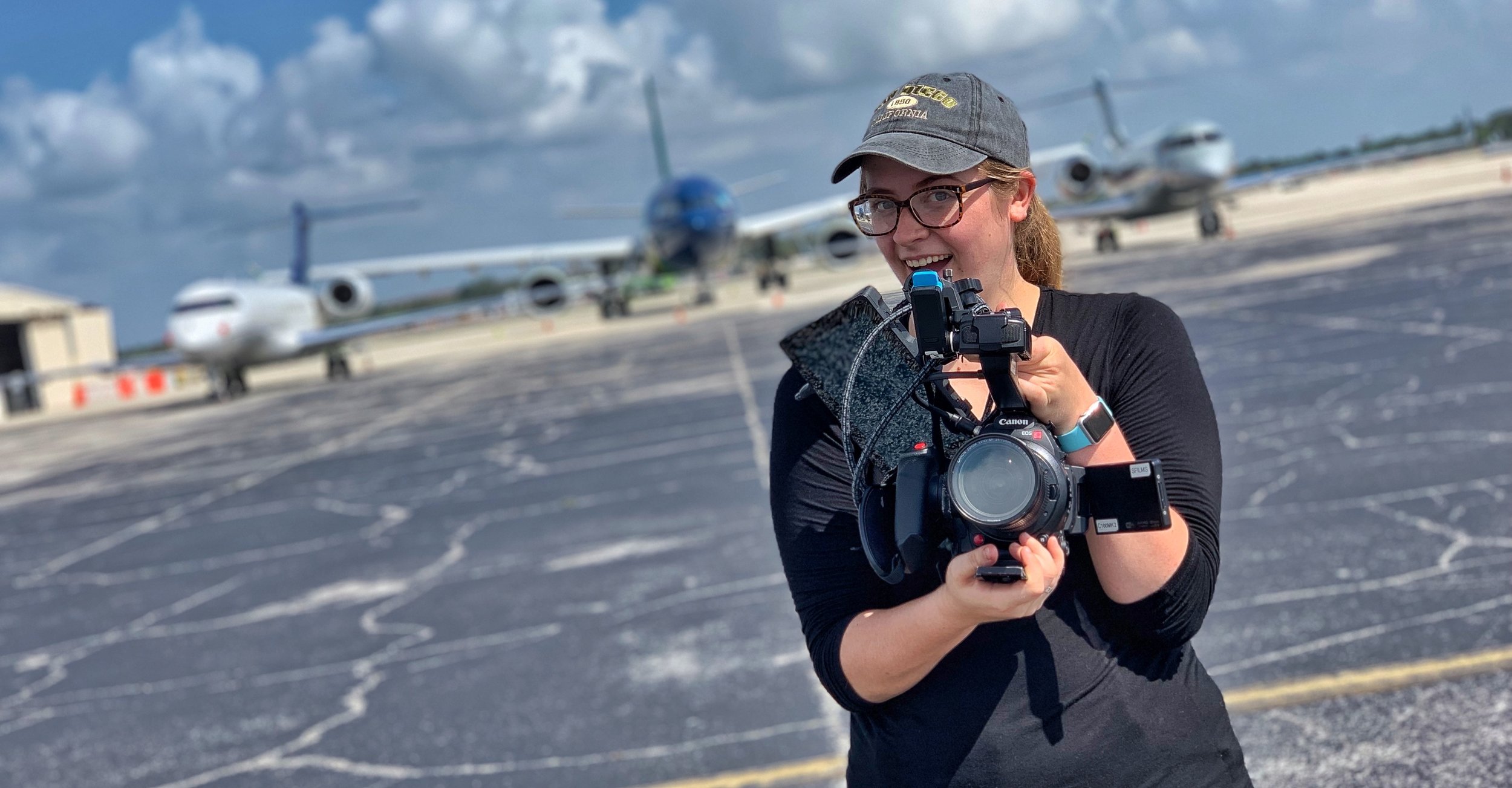 Young woman in black long-sleeve shirt holding cameras in an airport tarmac with airplanes in the background, smiling at the camera.