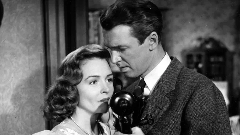 A black-and-white scene from an old movie showing a man and woman close to each other. The man is holding a vintage telephone receiver to his ear while the woman looks on with a serious expression. They are indoors, with a blurred background.