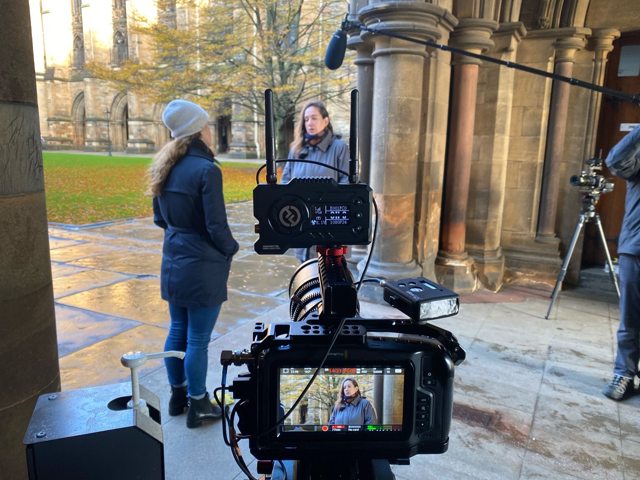 A behind-the-scenes view of a film or video shoot outside a historic building with stone columns and gothic architecture. Two women in jackets and hats are engaged in conversation, with a professional camera setup aimed at them, and a person operatin