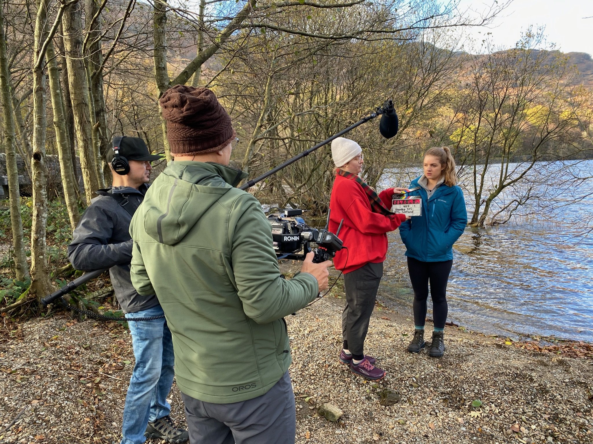 Filming crew is shooting a scene with two women near a lake in a wooded area during fall. One woman in a red jacket is speaking while holding a clapperboard, and the other woman in a blue jacket listens. The crew includes a cameraman with a camera mo