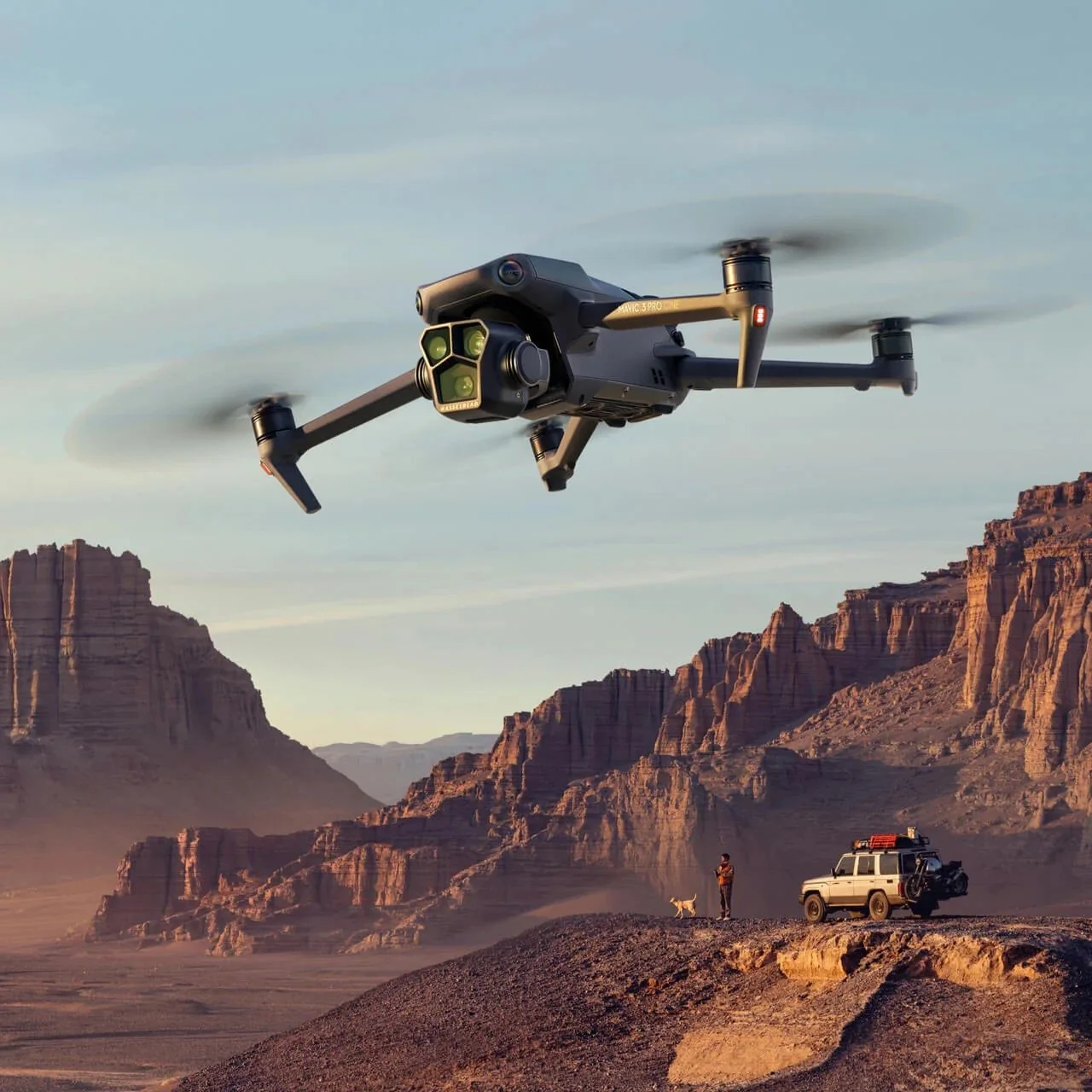 A drone flying over a desert landscape with rocky cliffs, a person standing with a dog near a parked vehicle, and the vast canyon in the background.