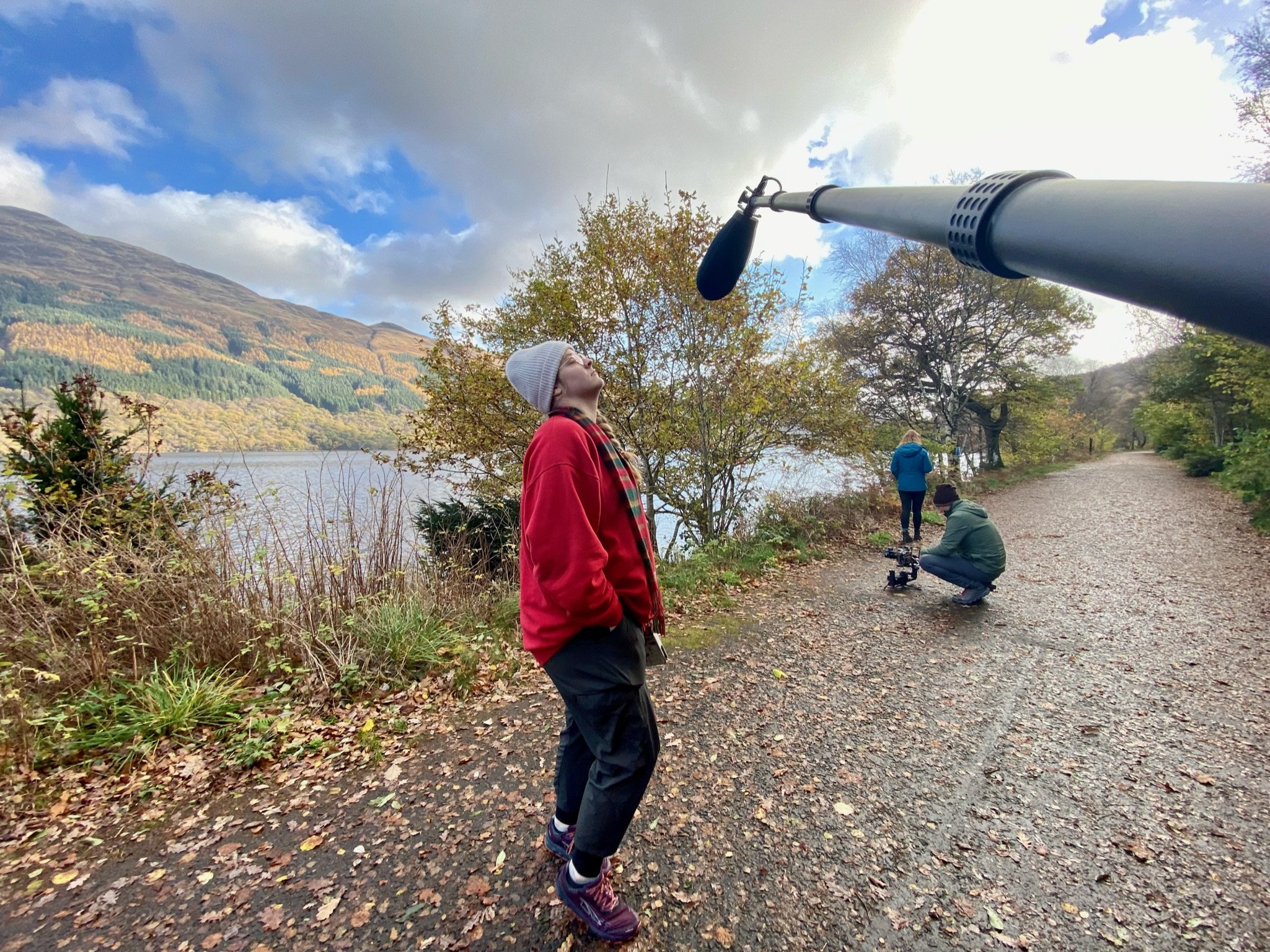 A woman stands outdoors near a lake, looking up at the sky during the daytime. Three people, including a man operating a camera, are nearby. The scene is set on a wooded trail with falls foliage in the background, and the sky is partly cloudy.