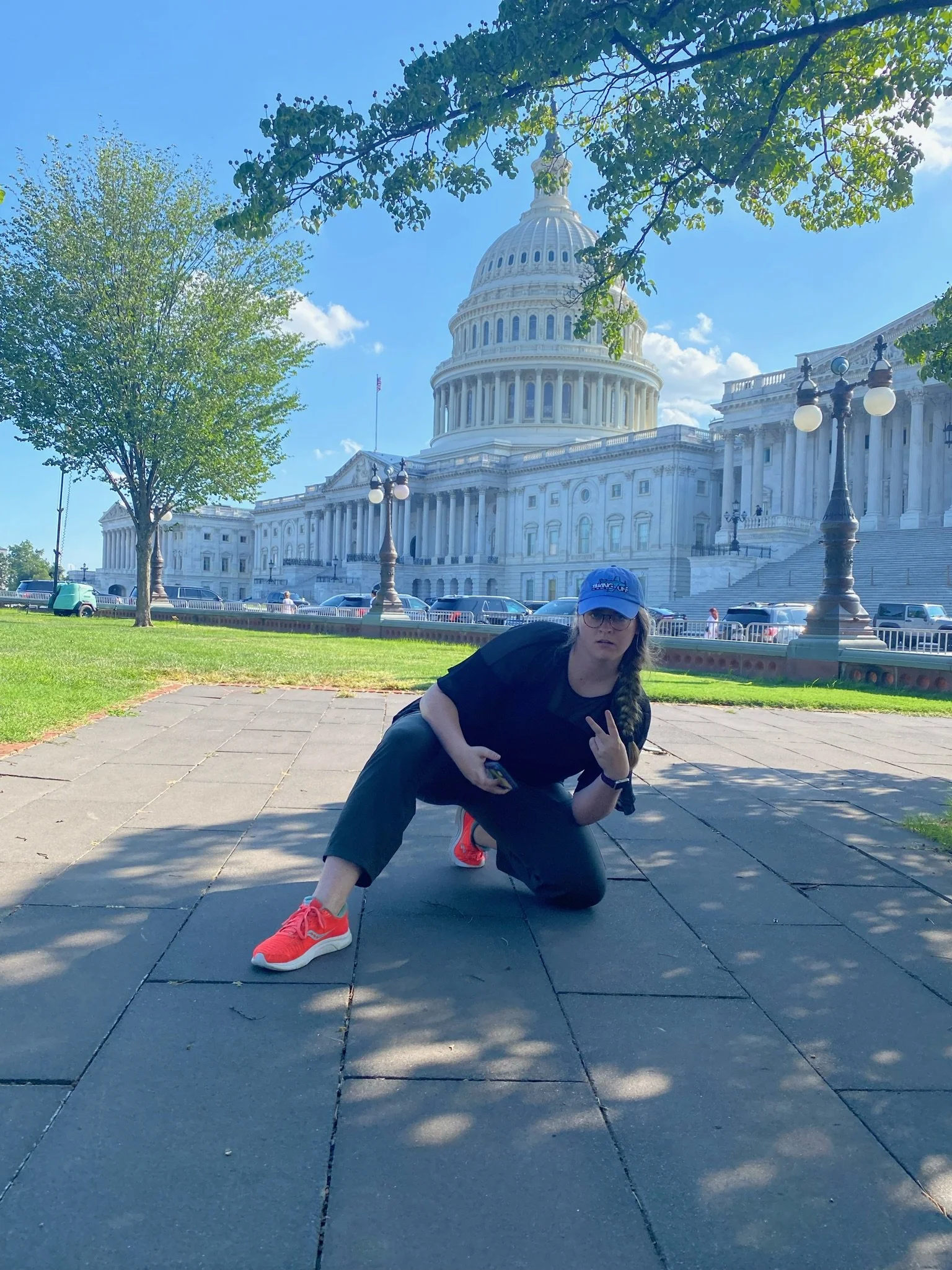 A young woman crouching on a sidewalk in front of the U.S. Capitol building in Washington, D.C., making a peace sign with her hand, wearing a blue cap, black shirt, gray pants, and red shoes, with trees and parked cars in the background on a sunny da