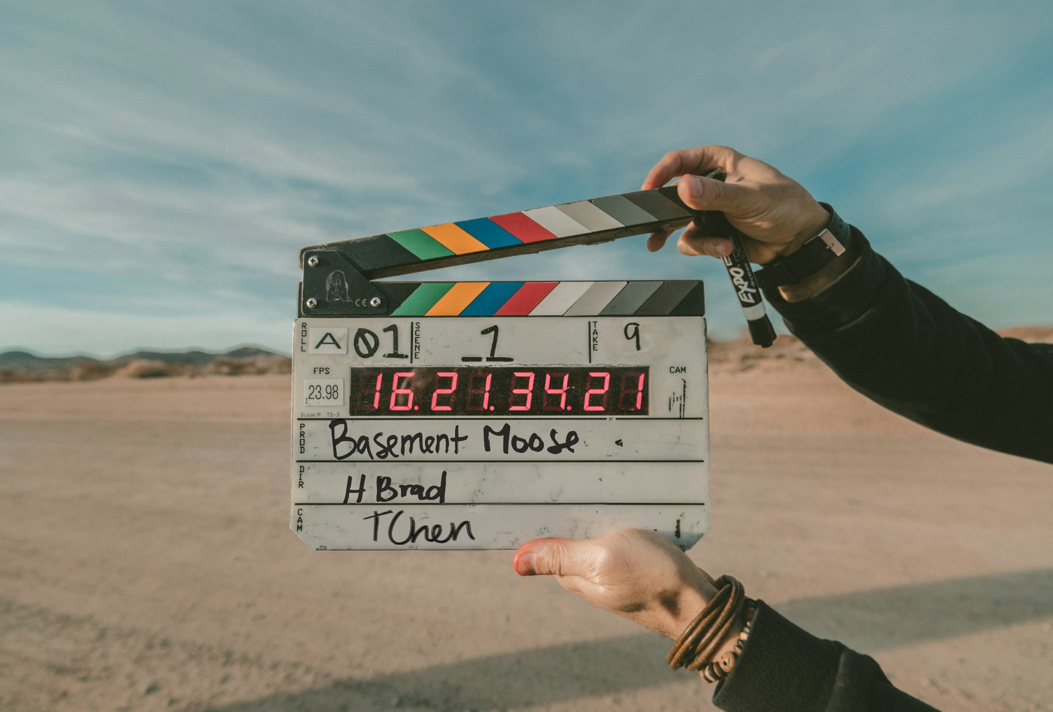 Clapperboard held in a desert with blue sky background, displaying scene information, handwritten notes, and a digital timestamp.