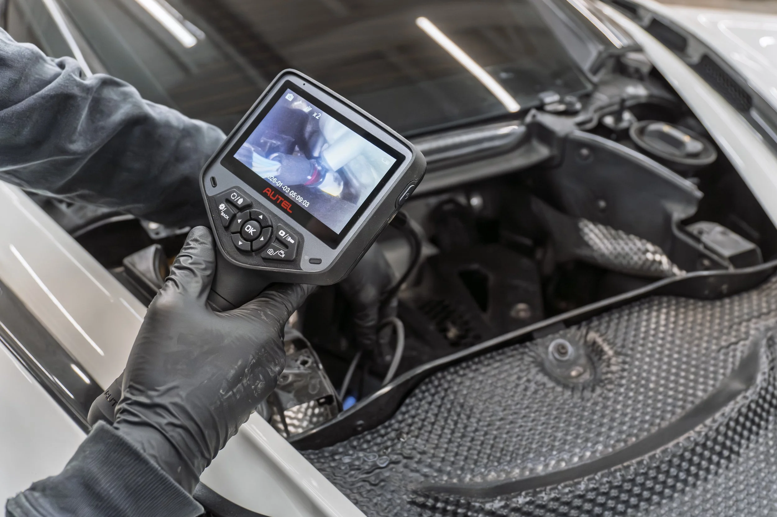 Close-up of a person wearing black gloves holding a diagnostic tool or camera, inspecting the engine bay of a car, with the engine exposed and a carbon fiber hood visible.