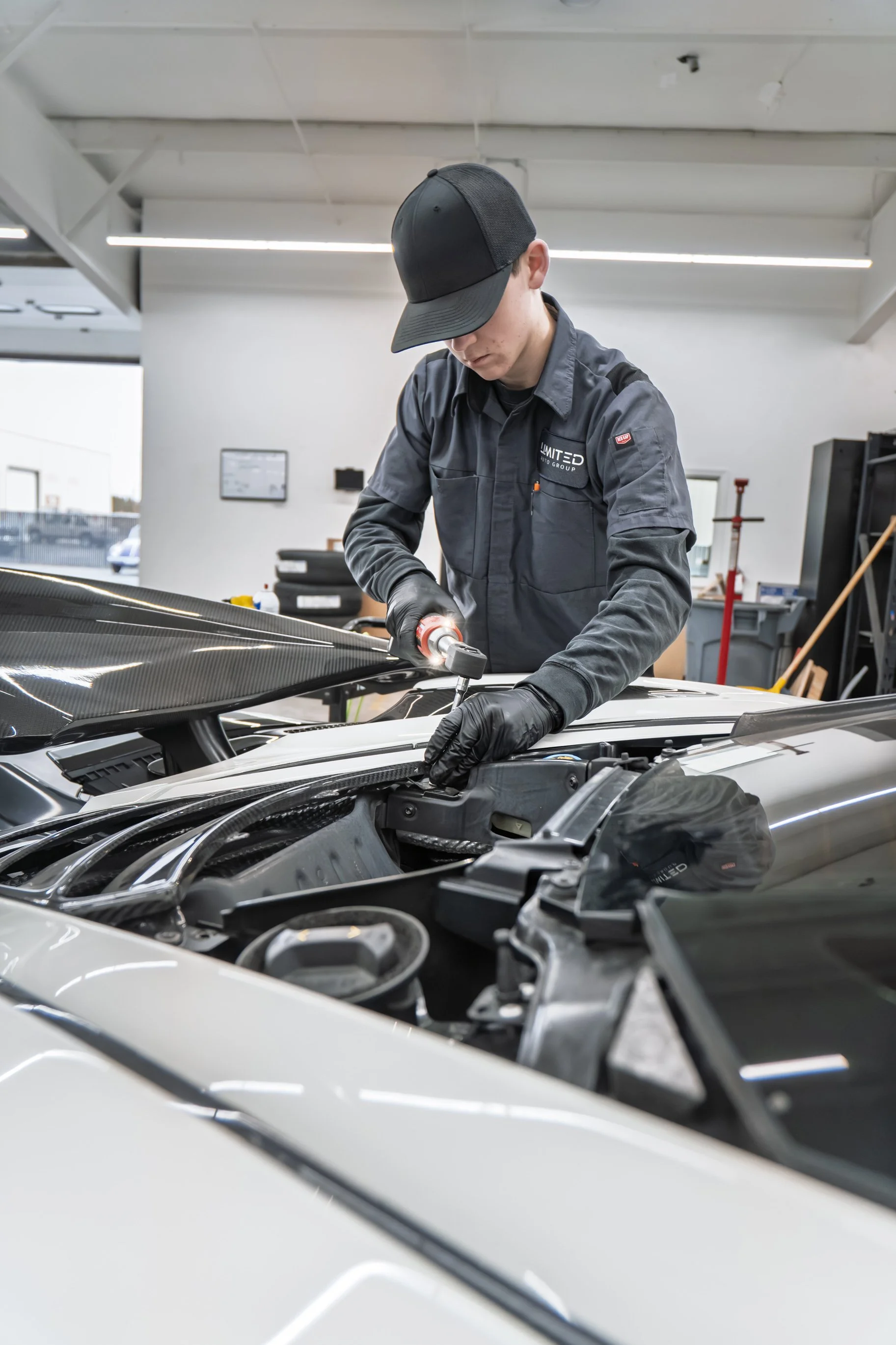 A person wearing a black cap, gray work shirt, and gloves is working on a car engine in a well-lit workshop.
