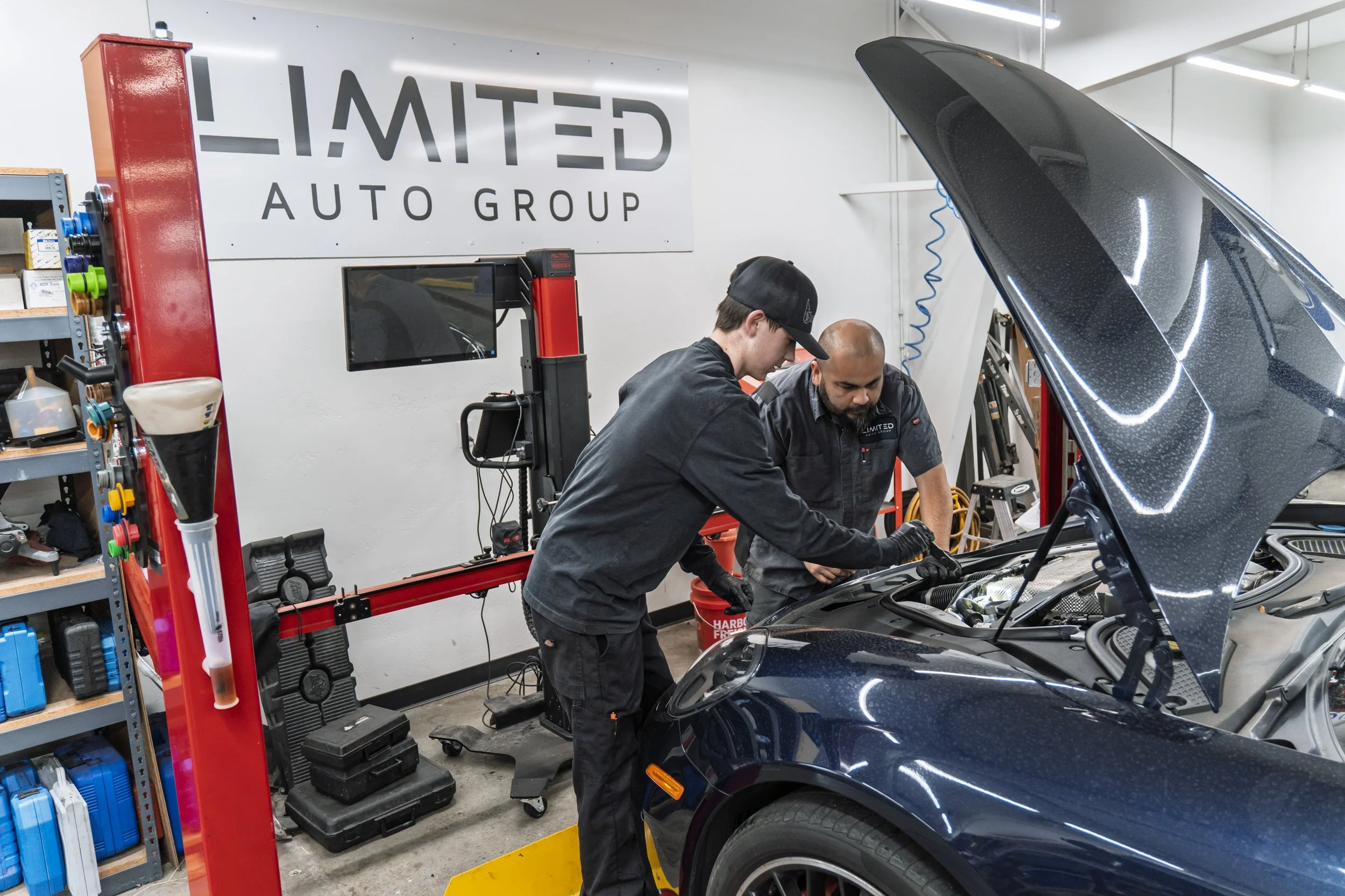 Two mechanics working on a dark blue car in an auto repair shop. One is under the hood inspecting or repairing, while the other looks on. The shop has a white wall with a large sign that reads 'LIMITED AUTO GROUP' and various shelves with tools and supplies.