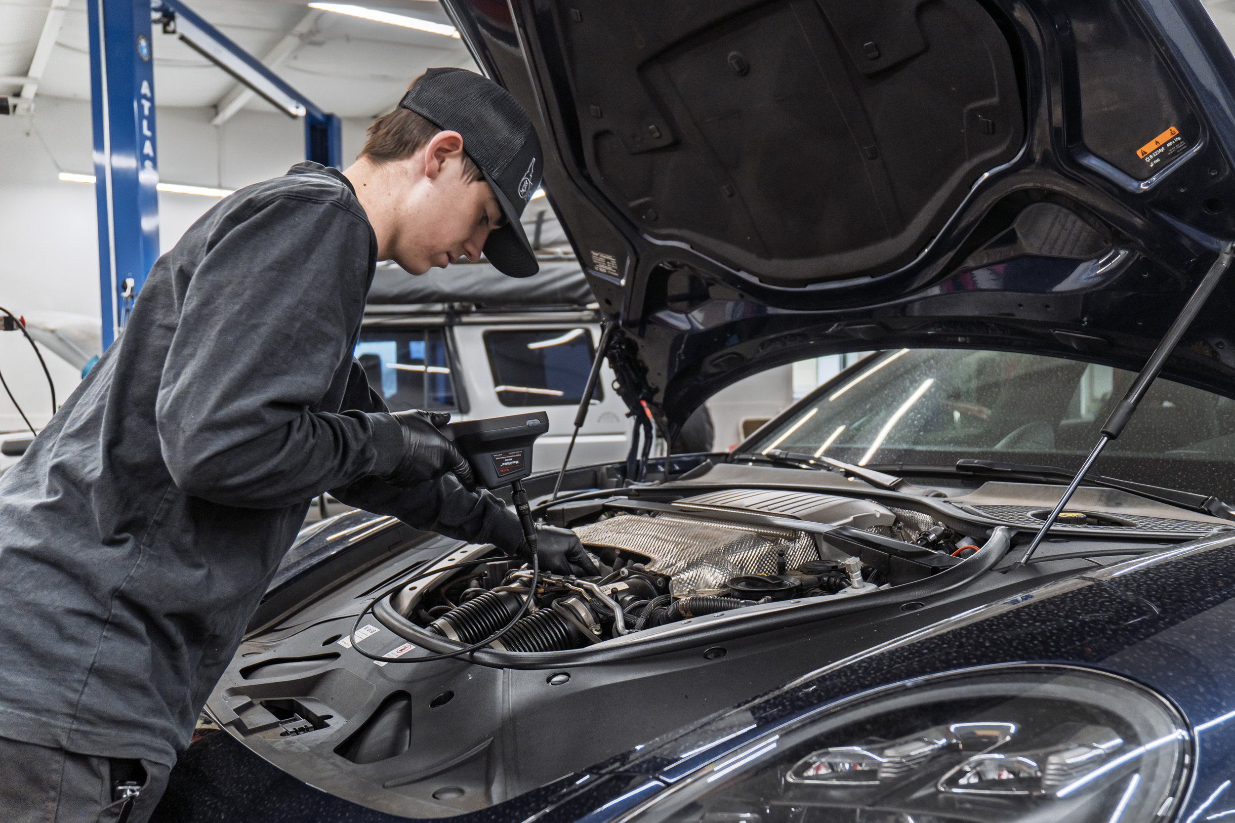 A young mechanic wearing a black cap, gray jacket, and gloves inspects the engine of a black car in an auto repair shop.